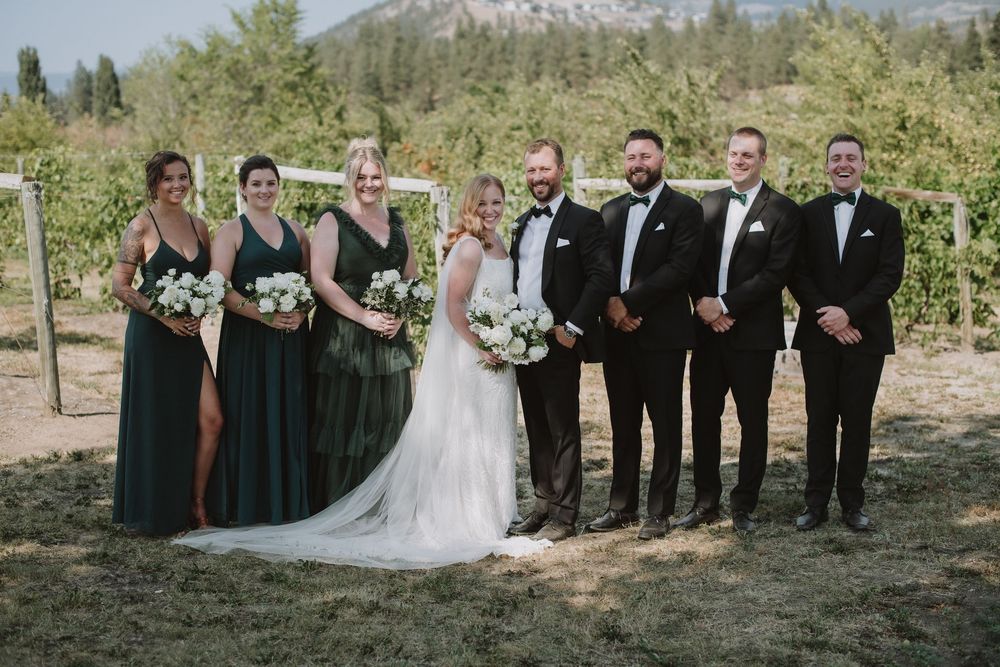 Wedding party poses in vineyard, bridesmaids in emerald gowns and groomsmen in black tuxedos flanking bride and groom.