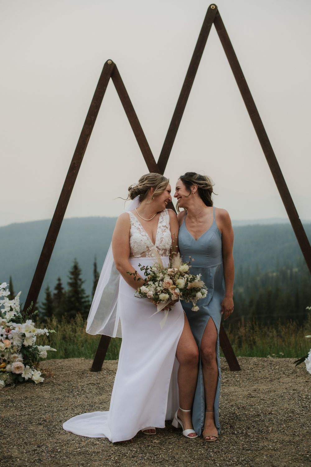 Two brides share a romantic moment at an outdoor mountain wedding ceremony beneath a geometric wooden arch.