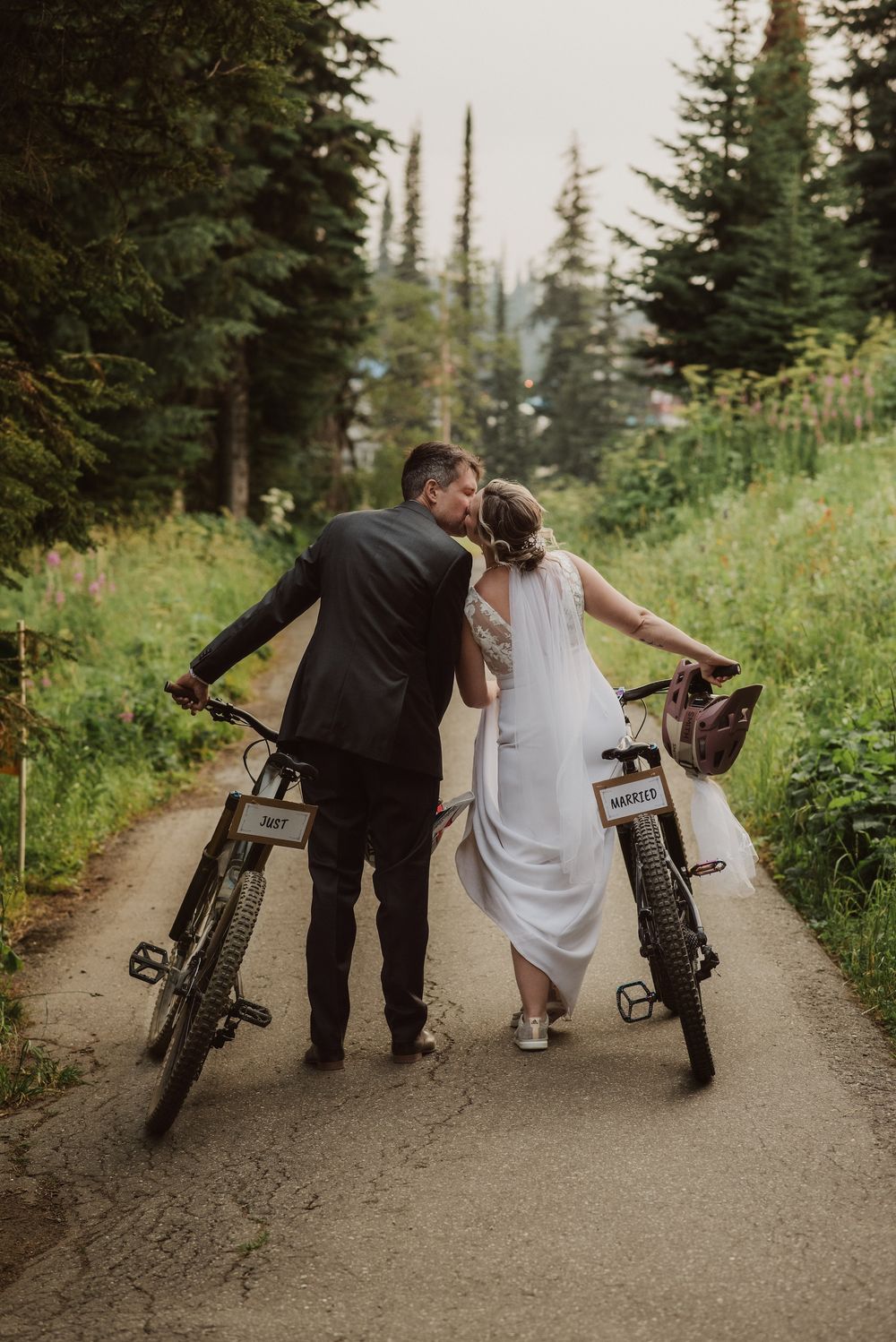 Newlyweds kiss on a forest trail with their bicycles labeled Mr. and Mrs. during a scenic outdoor wedding celebration.