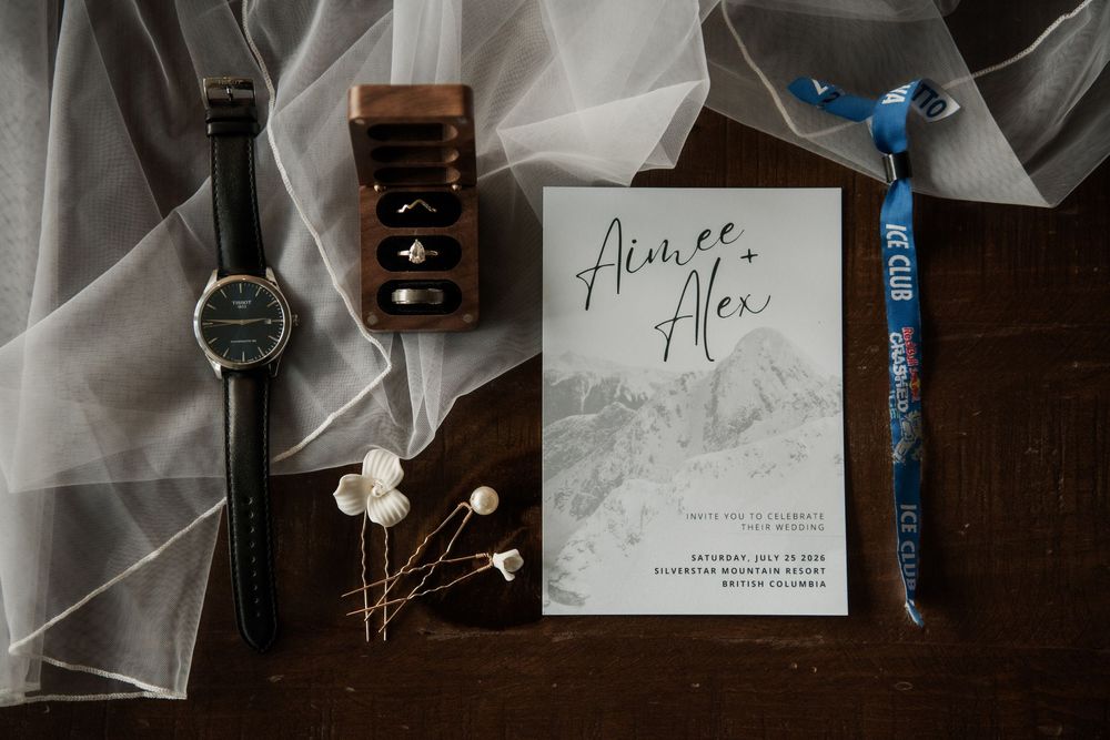 Wedding flat lay with invitation for Aimee and Alex, a watch, ring box, and blue wristband on dark wood.