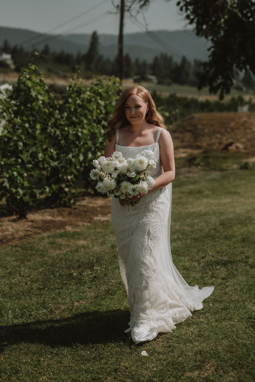 Bride in lace wedding gown holding white floral bouquet standing on green lawn with mountain backdrop.