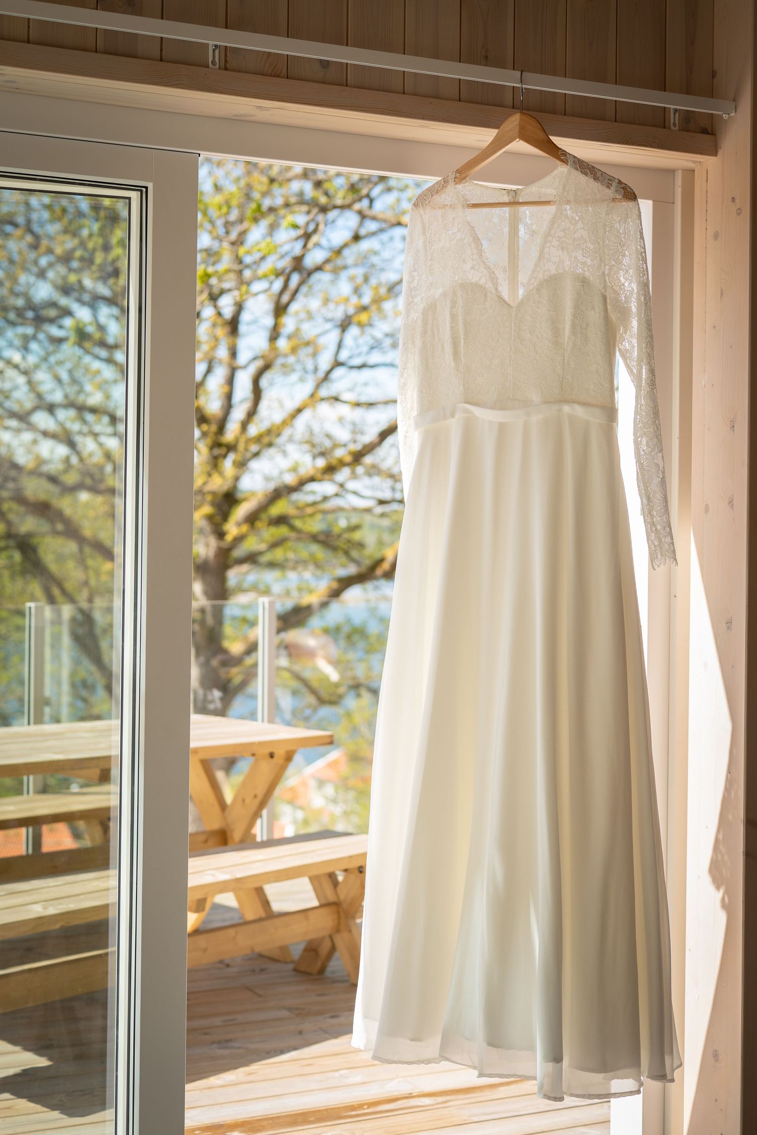 Elegant white lace wedding dress with long sleeves hanging on wooden hanger in sunlit doorway with nature backdrop.
