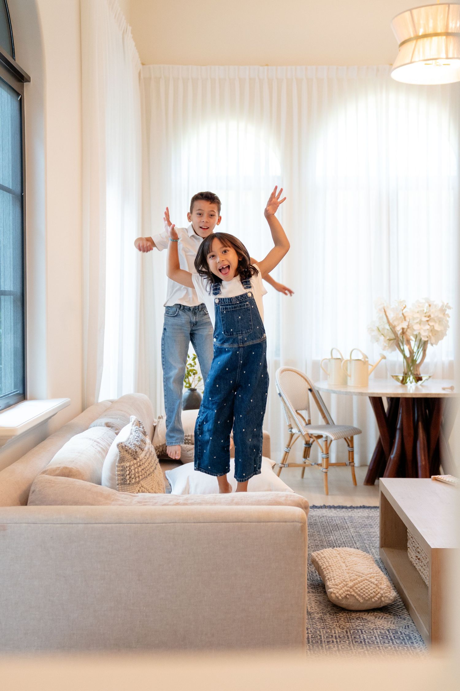 Two children joyfully jumping on a couch in a bright modern living room, wearing jeans and white tops.