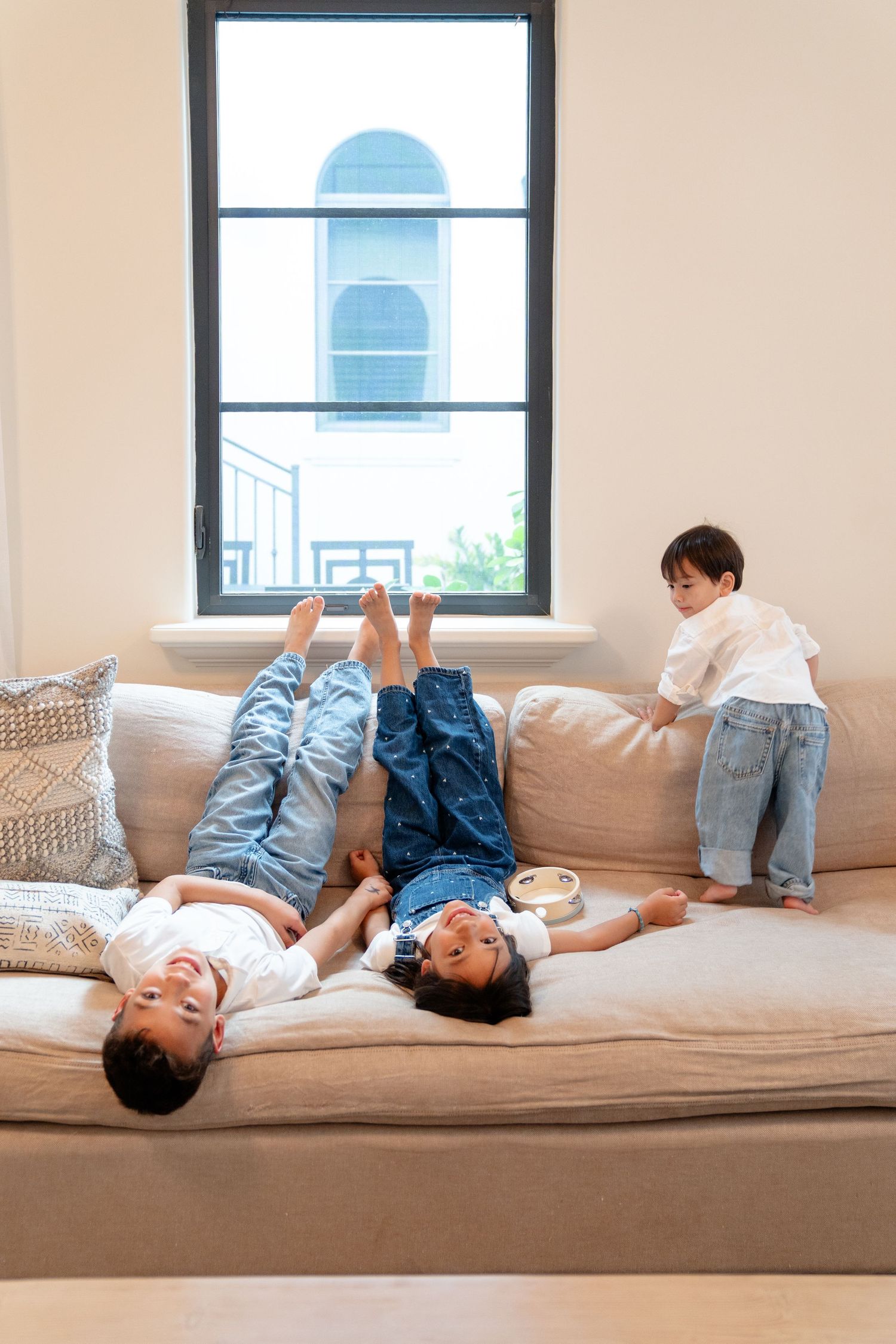 Family relaxing on a beige sofa, two children lying down laughing while a toddler stands by the window.