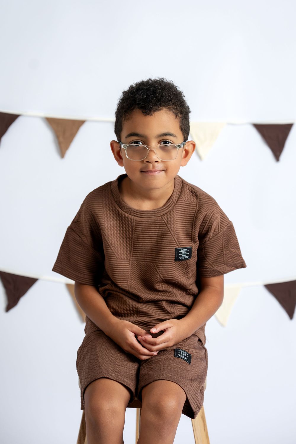 A young boy with glasses wearing a brown textured outfit sits smiling in front of party bunting.