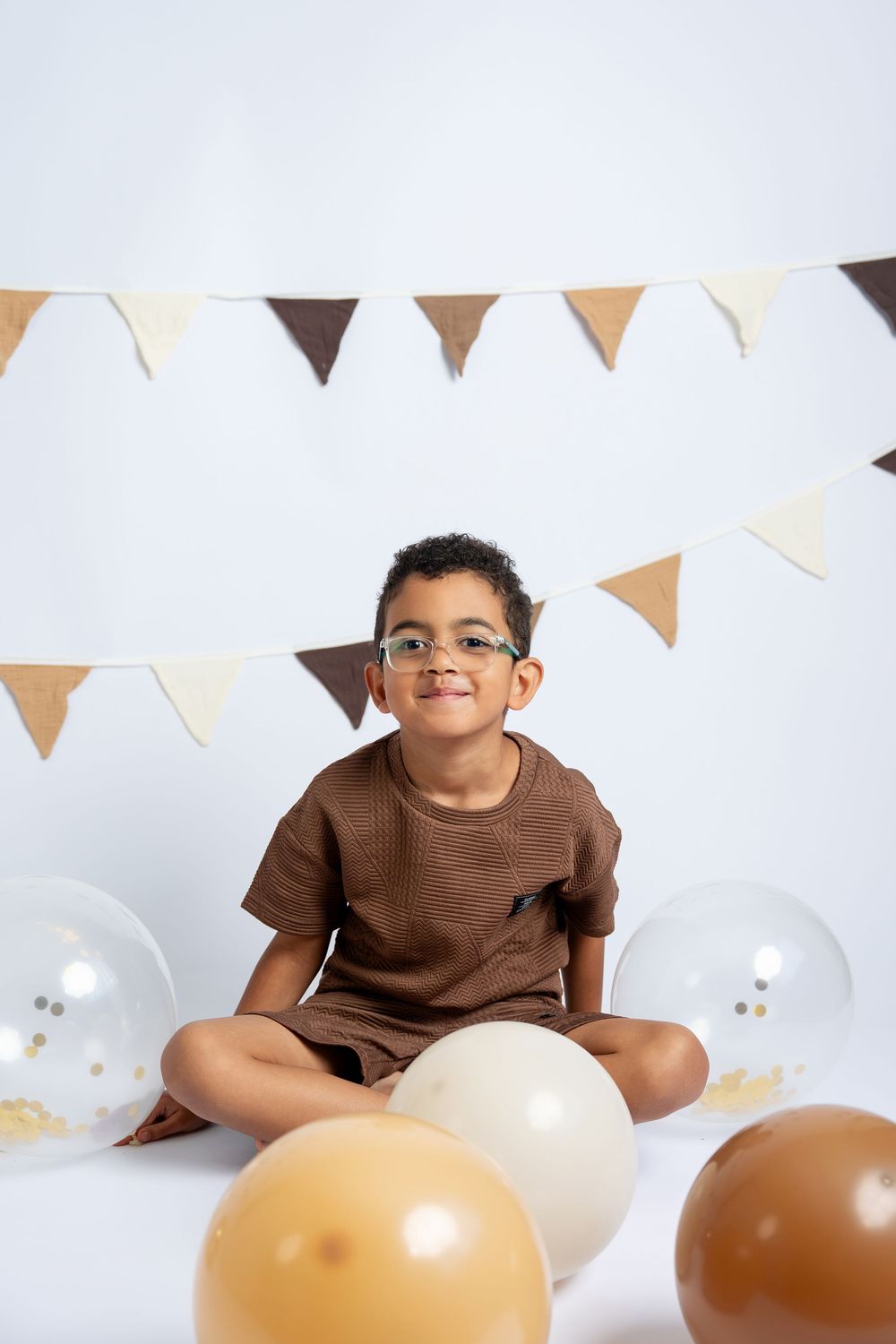 A young boy wearing glasses and a brown outfit sits cross-legged among balloons at a birthday celebration.