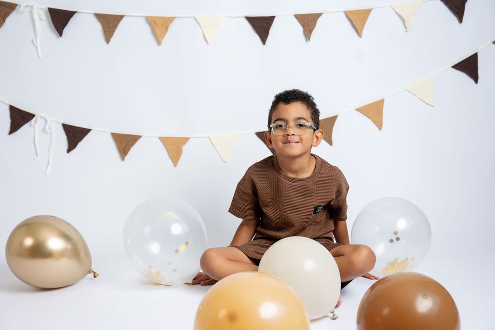 Smiling boy in glasses and brown shirt sits cross-legged with balloons and bunting at a birthday celebration.