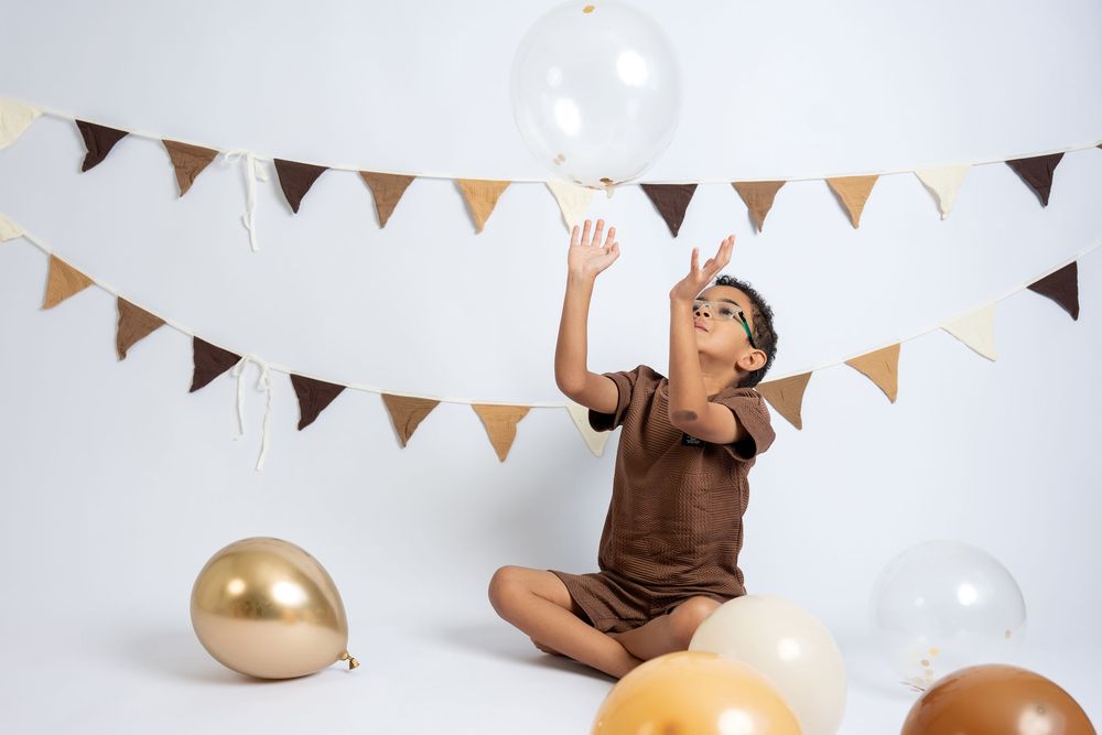 A joyful boy sitting cross-legged among gold and clear balloons, reaching up playfully under brown and gold party bunting.