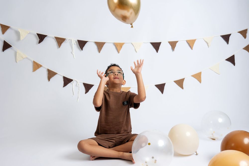 A boy in glasses sits cross-legged reaching up toward a gold balloon at a birthday party with bunting decorations.