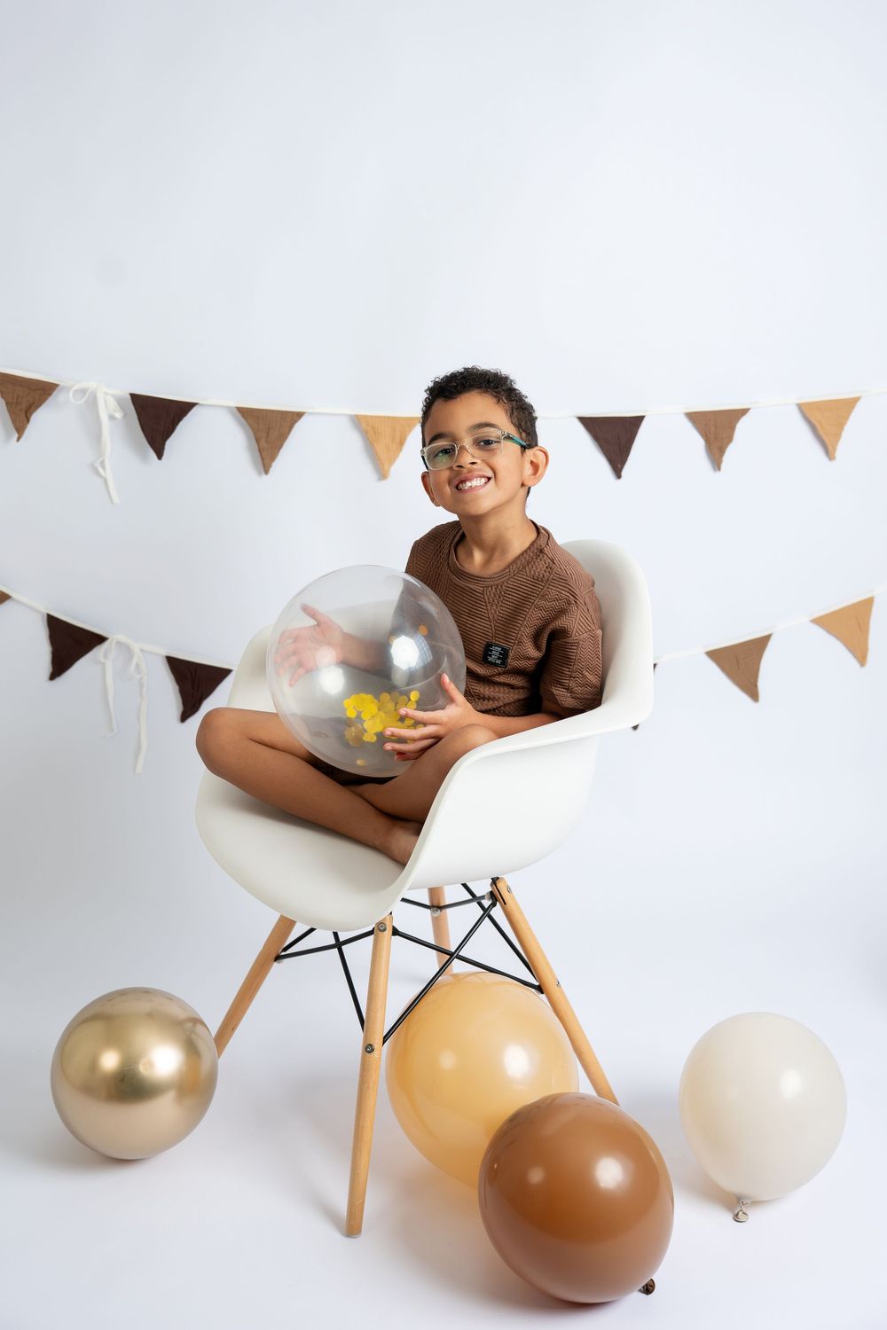 Smiling boy with glasses sits on white chair holding clear balloon, surrounded by gold and brown balloons and bunting.