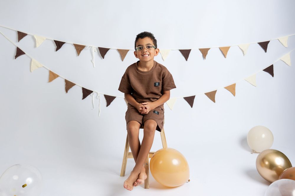 Boy in brown outfit sits on stool with gold and white balloons and triangle bunting backdrop.