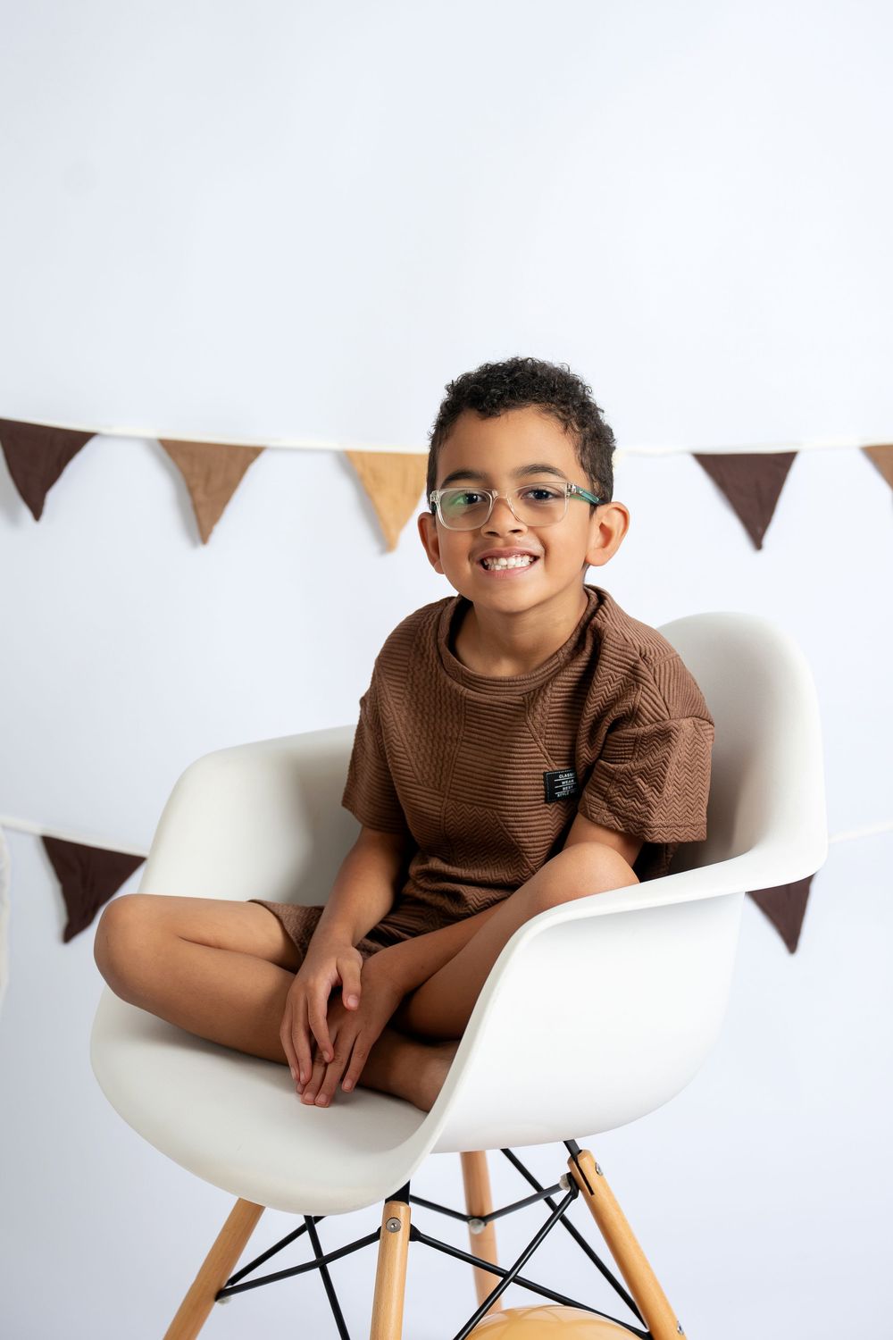 Smiling boy in brown outfit and glasses sitting cross-legged on white chair with bunting backdrop.
