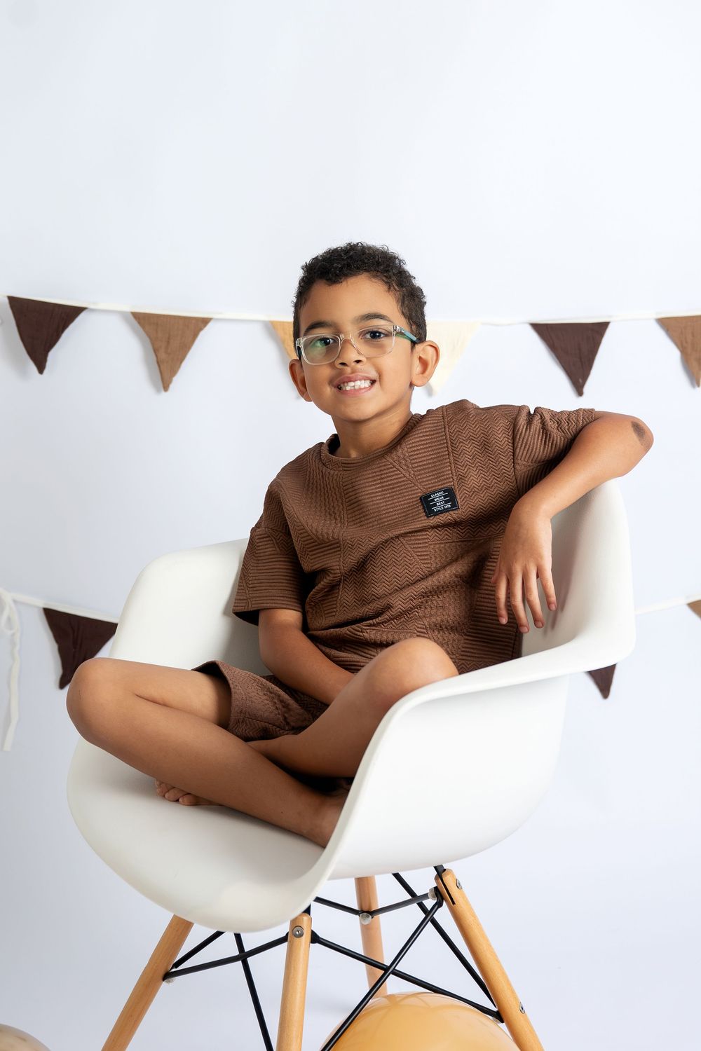 A smiling boy in brown outfit and glasses sits cross-legged on a white chair with decorative bunting behind him.