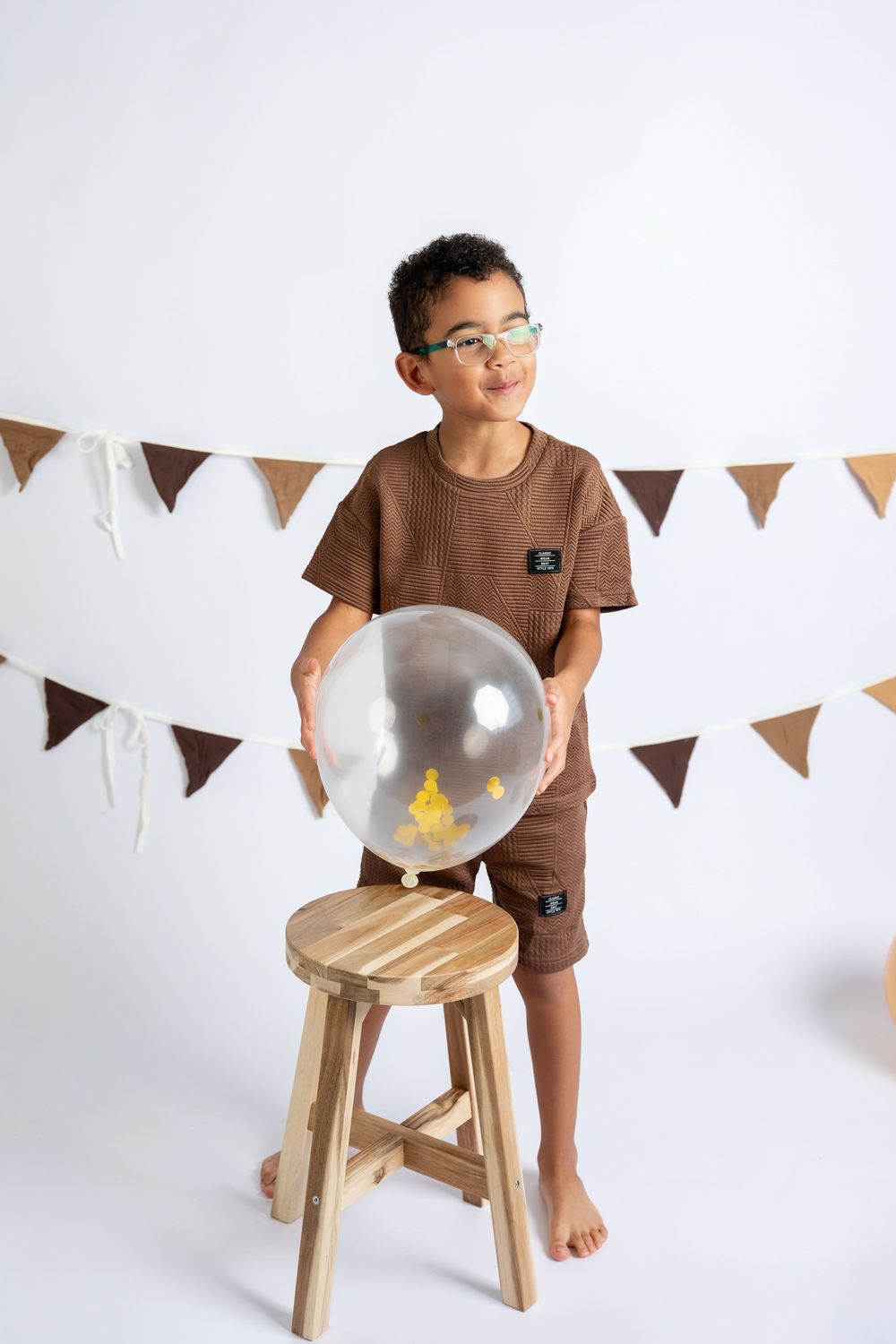 Young boy in brown outfit holding a clear balloon with yellow decor, standing by a wooden stool with bunting backdrop.