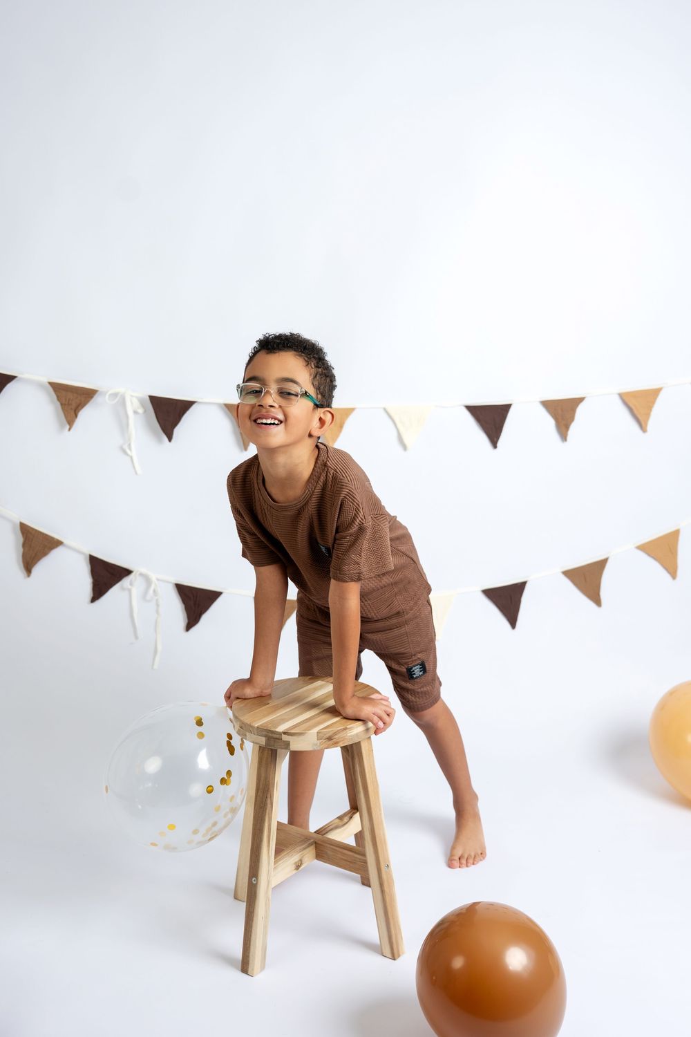 Happy boy in brown outfit and glasses leaning on wooden stool with balloons and bunting backdrop.