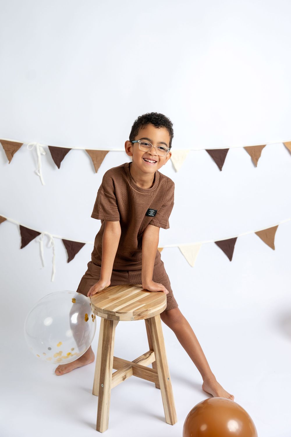 Smiling boy in brown outfit sitting on wooden stool with balloons and bunting backdrop in a white studio setting.