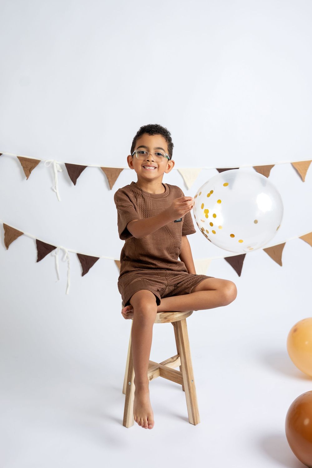 A smiling boy in brown outfit sits on stool holding clear balloon with gold confetti at birthday shoot.