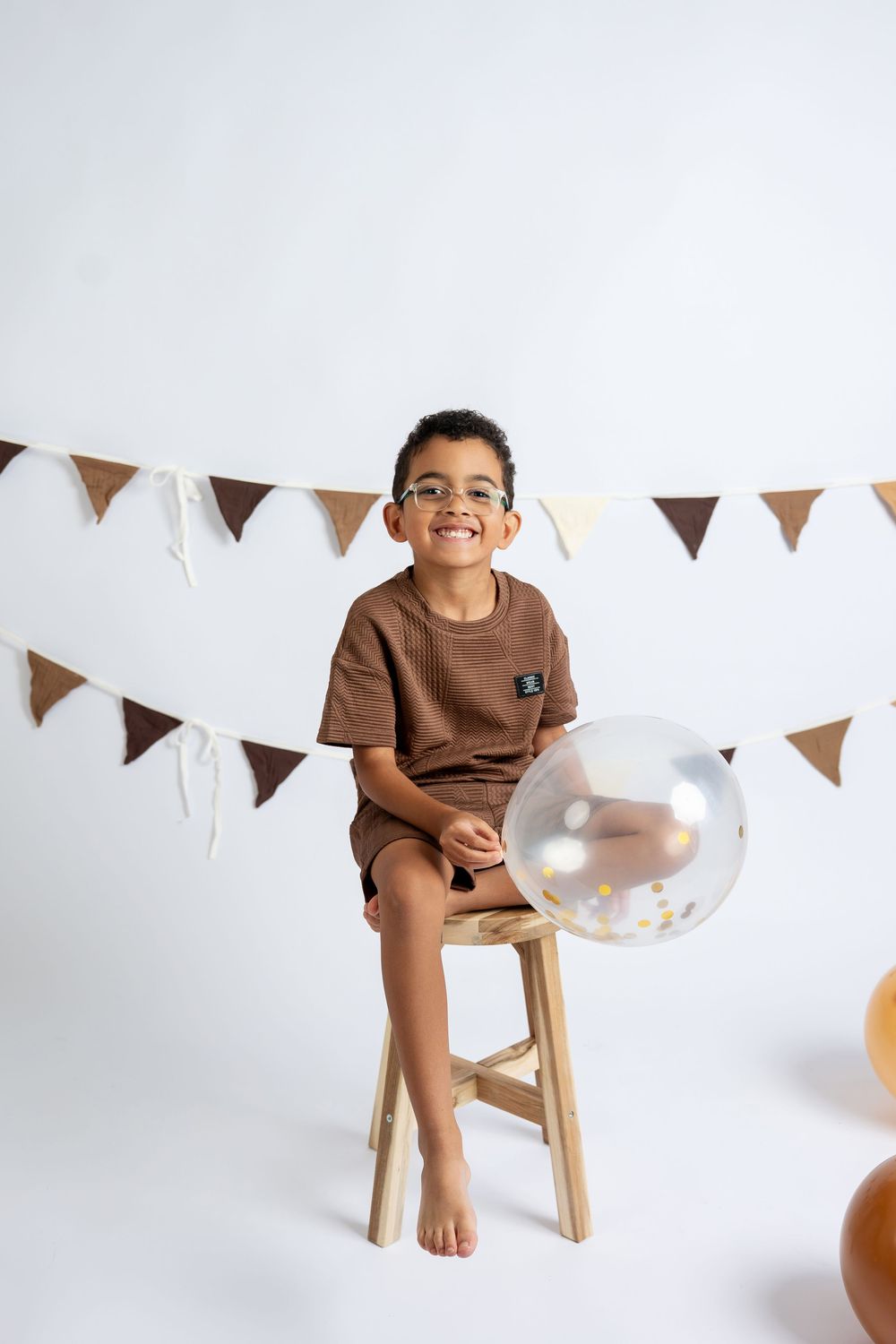 Smiling boy in brown outfit sits on wooden stool with clear balloon and bunting decorations on white background.