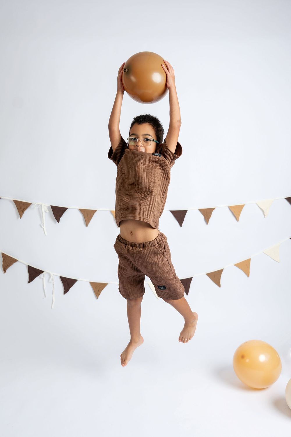 A young boy in glasses and brown outfit jumps holding an orange balloon, with festive bunting in the background.