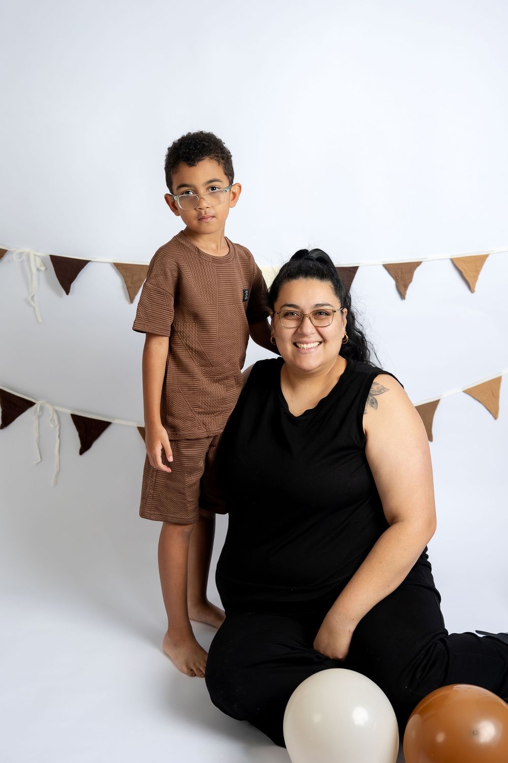A woman in black sits on the floor smiling beside a young boy in brown at a birthday party with bunting.