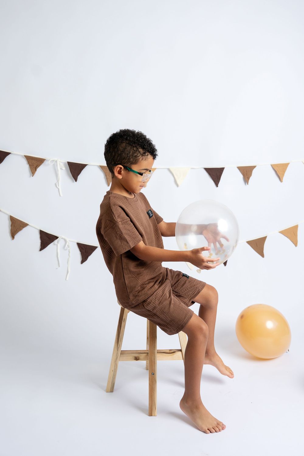 A young boy wearing glasses sits on a stool holding a clear balloon at a birthday party with festive bunting.