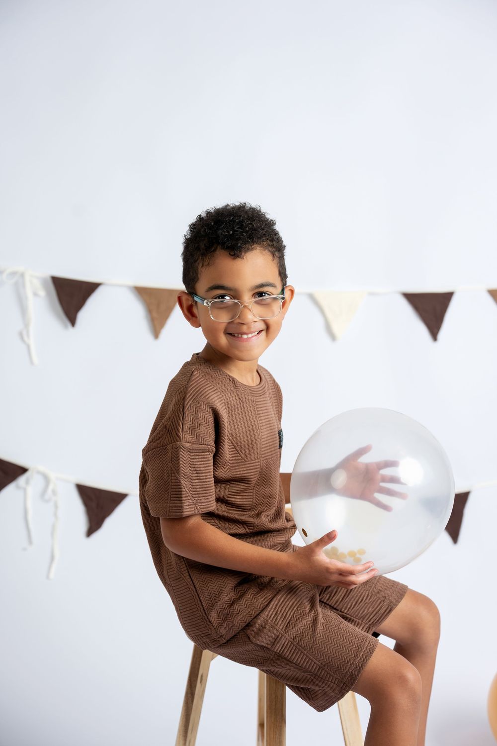 Smiling young boy wearing glasses and brown outfit sits on wooden stool holding clear balloon with party bunting backdrop.