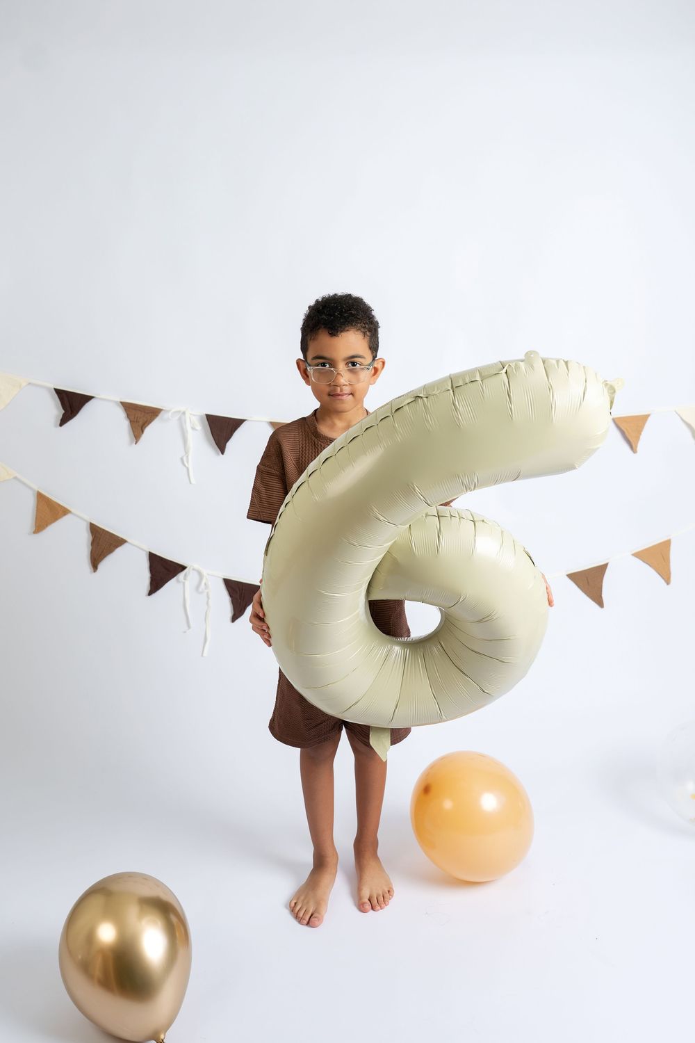 A young boy wearing glasses and brown outfit holds a large number 6 balloon at his birthday party.