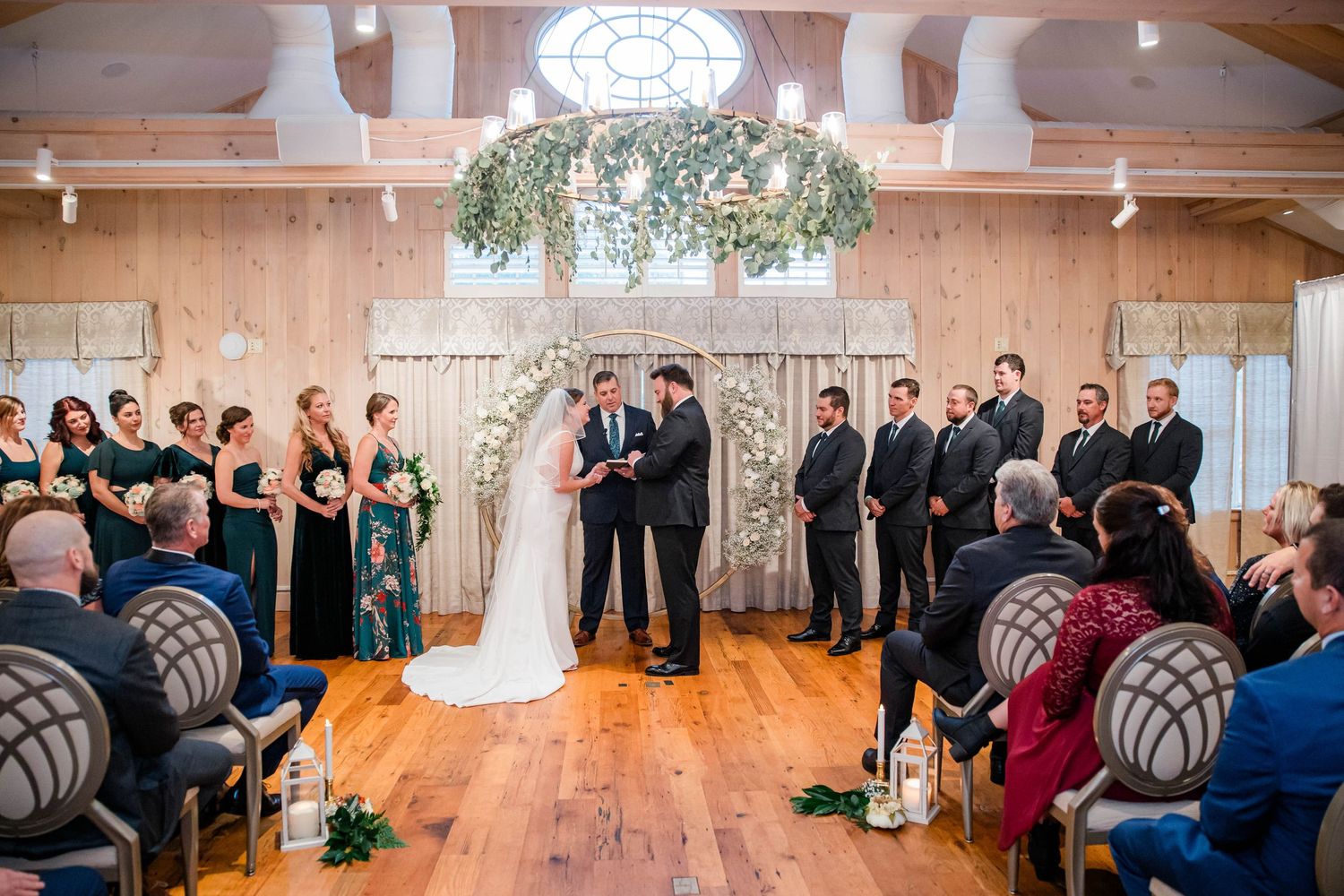 Elegant indoor wedding ceremony with floral arch, greenery chandelier, bridal party in emerald green, and guests seated on wooden chairs.