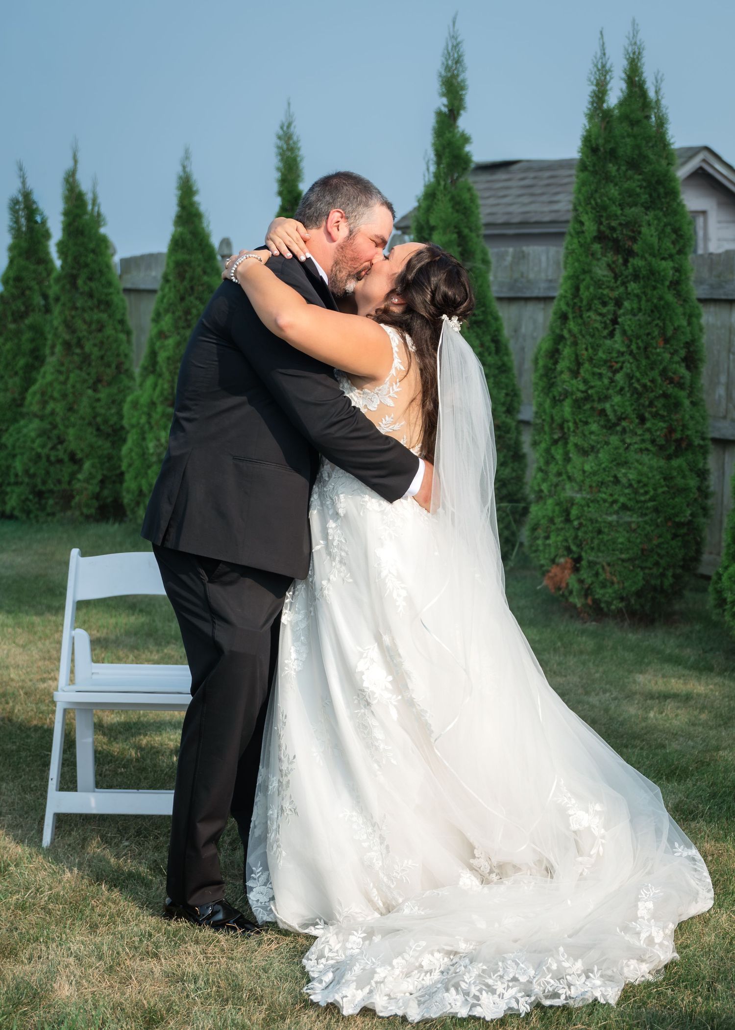Bride and groom share a romantic first kiss outdoors during their wedding ceremony, surrounded by tall green arborvitae trees.