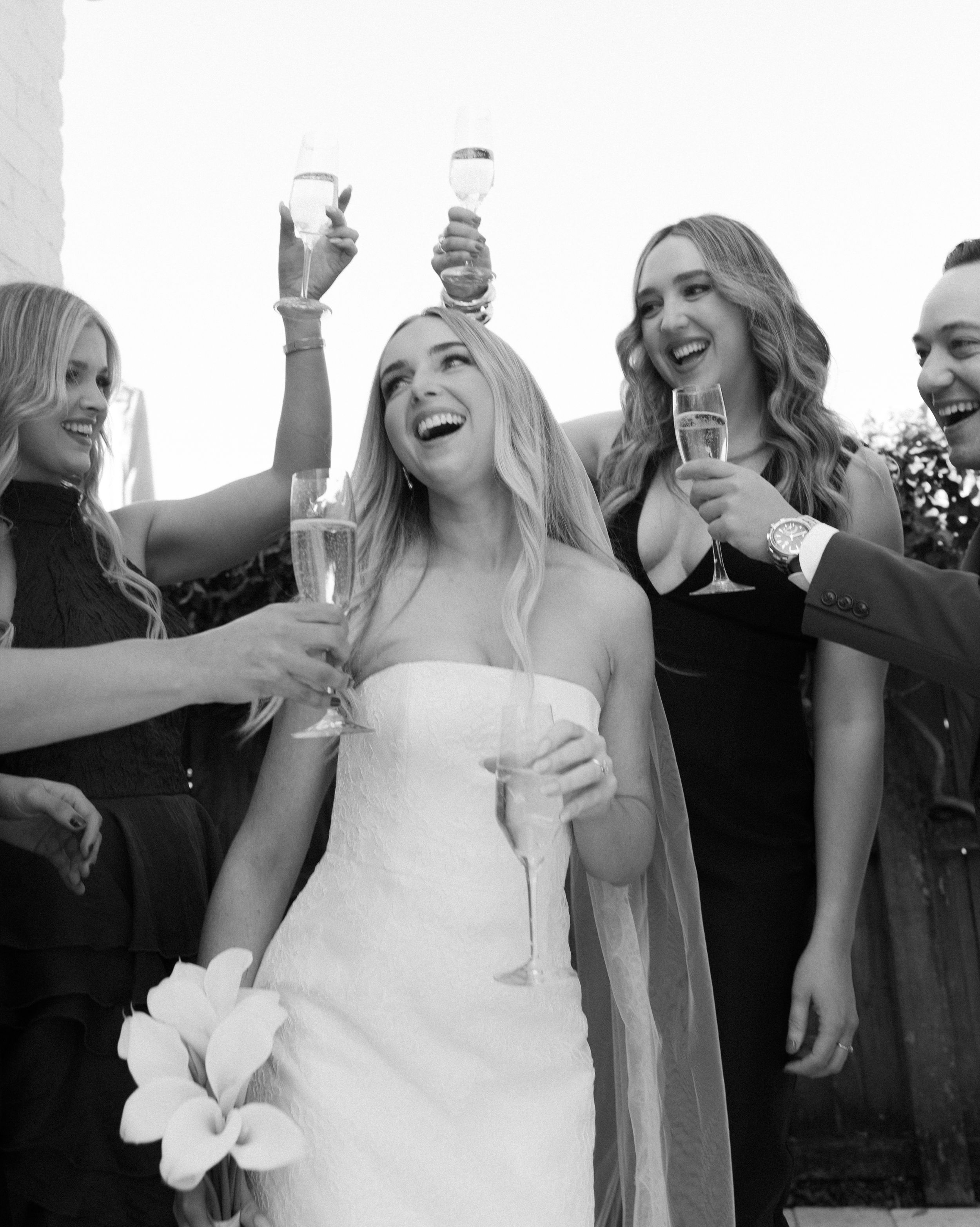 Black and white photo of a smiling bride in strapless gown toasting champagne with friends at a wedding celebration.
