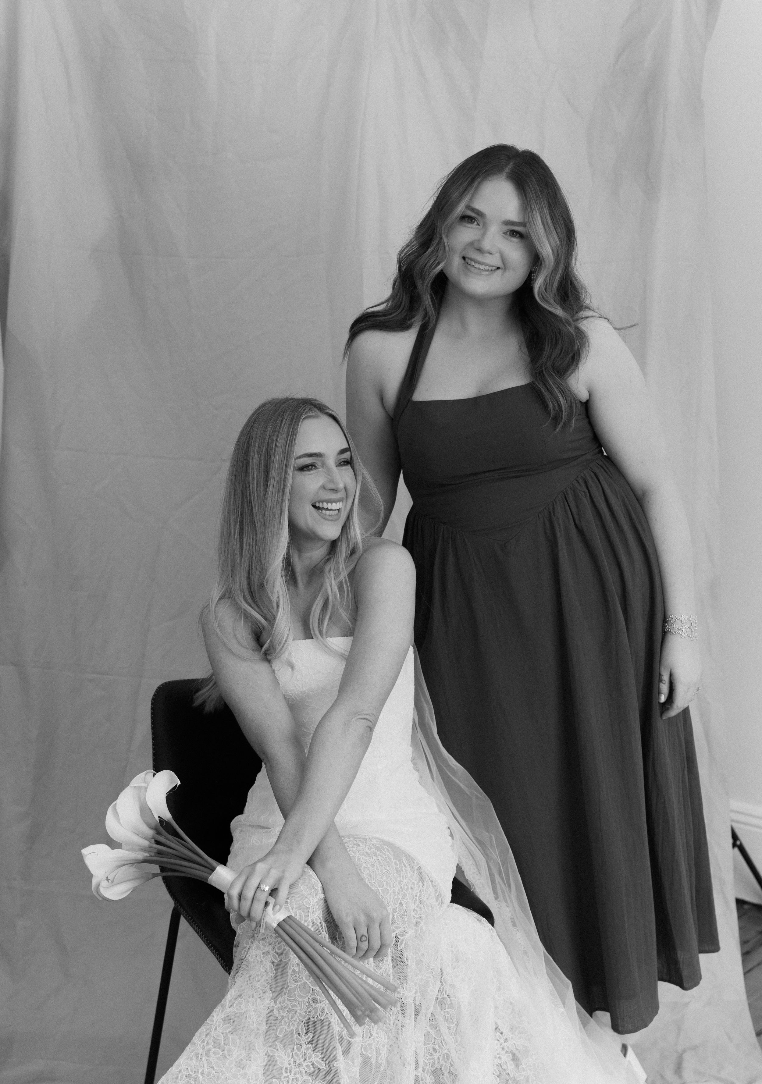 Black and white photo of a bride in a white gown holding flowers seated beside a bridesmaid in a dark halter dress.