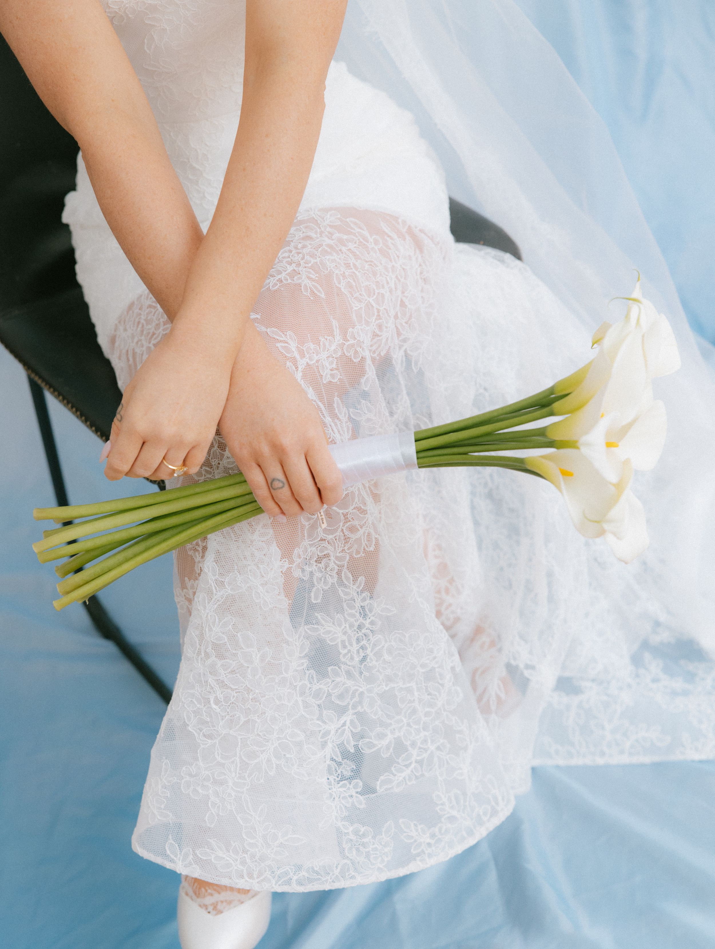 Bride in lace wedding dress holding white calla lily bouquet with blue veil draped over chair.