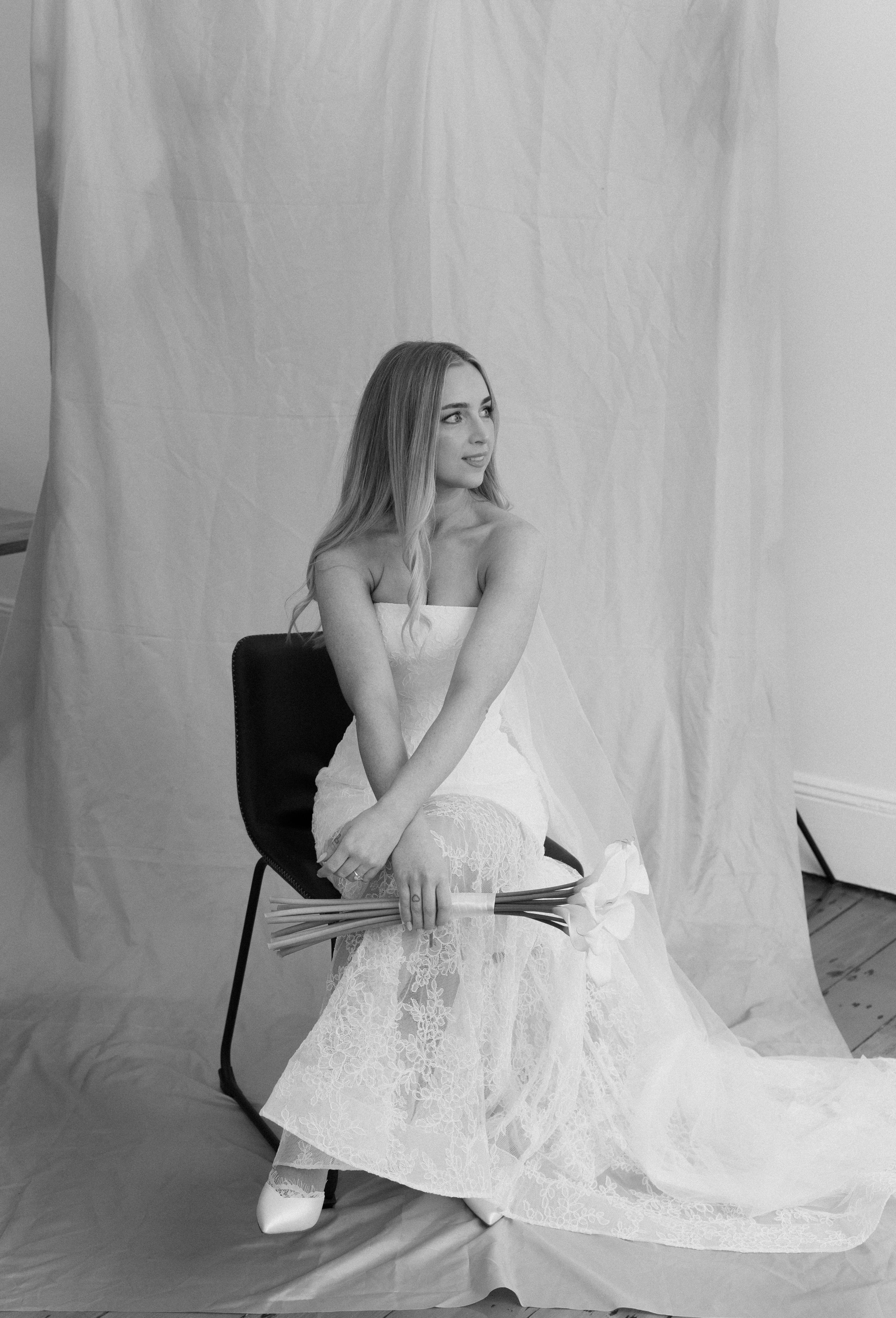Black and white photo of a woman in a white strapless lace wedding dress seated on a chair holding flowers.