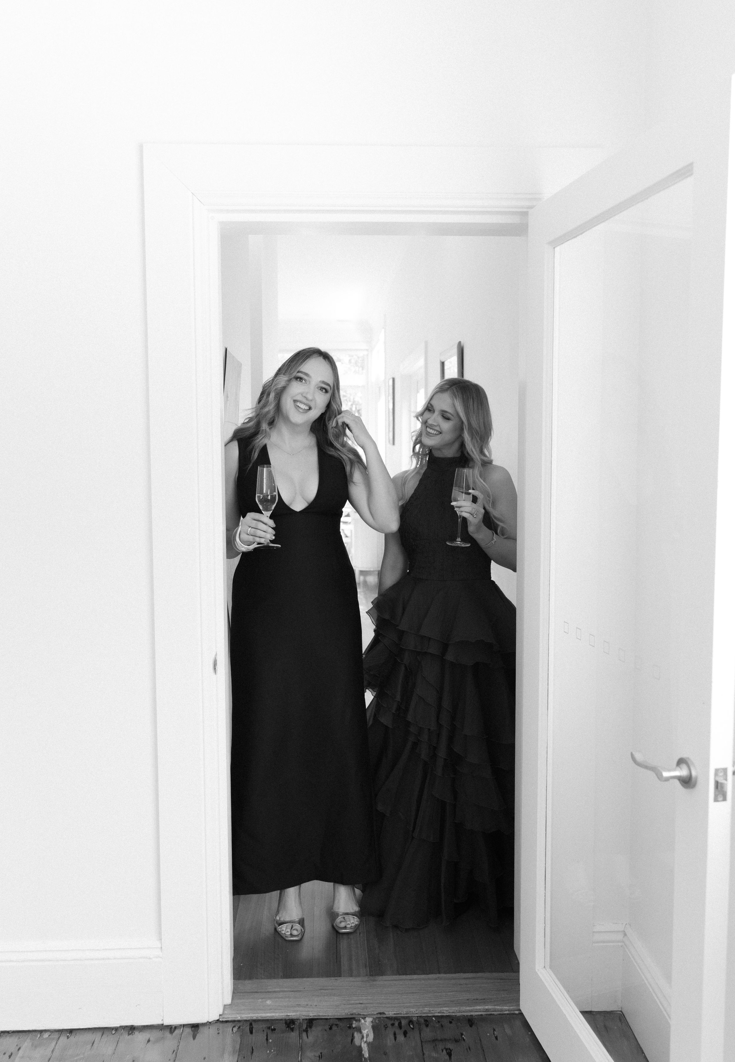 Two women in elegant black gowns holding champagne glasses pose in a doorway in black and white photo.