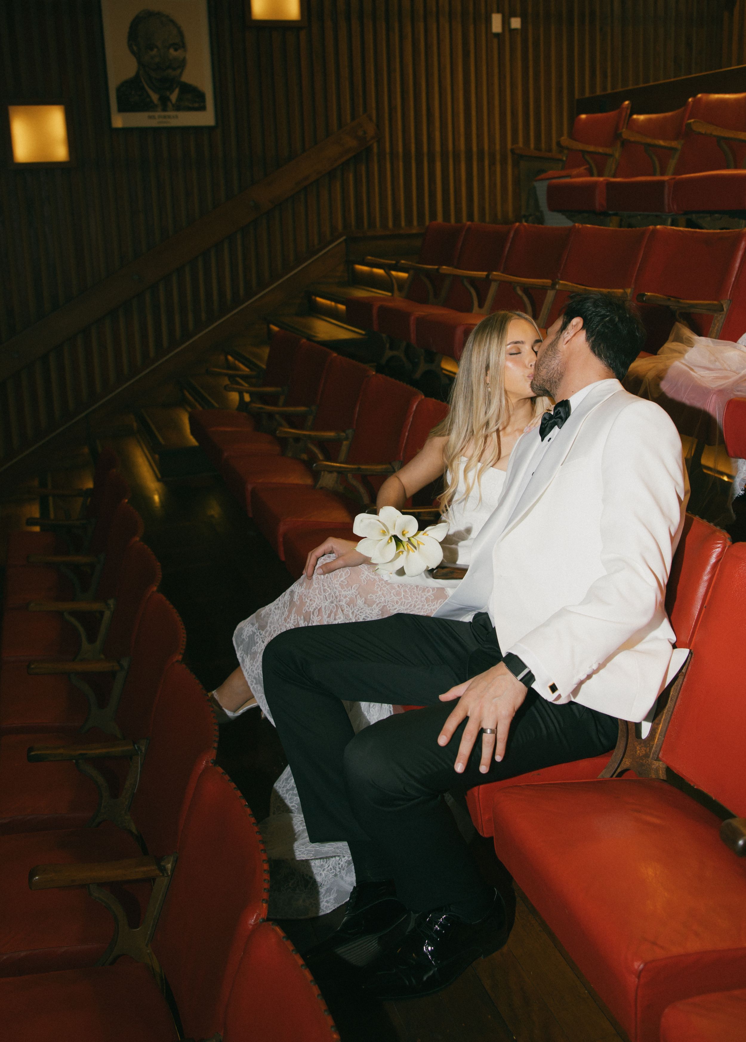 Couple in wedding attire kissing in red theater seats, groom in white tuxedo, bride holding white flowers.