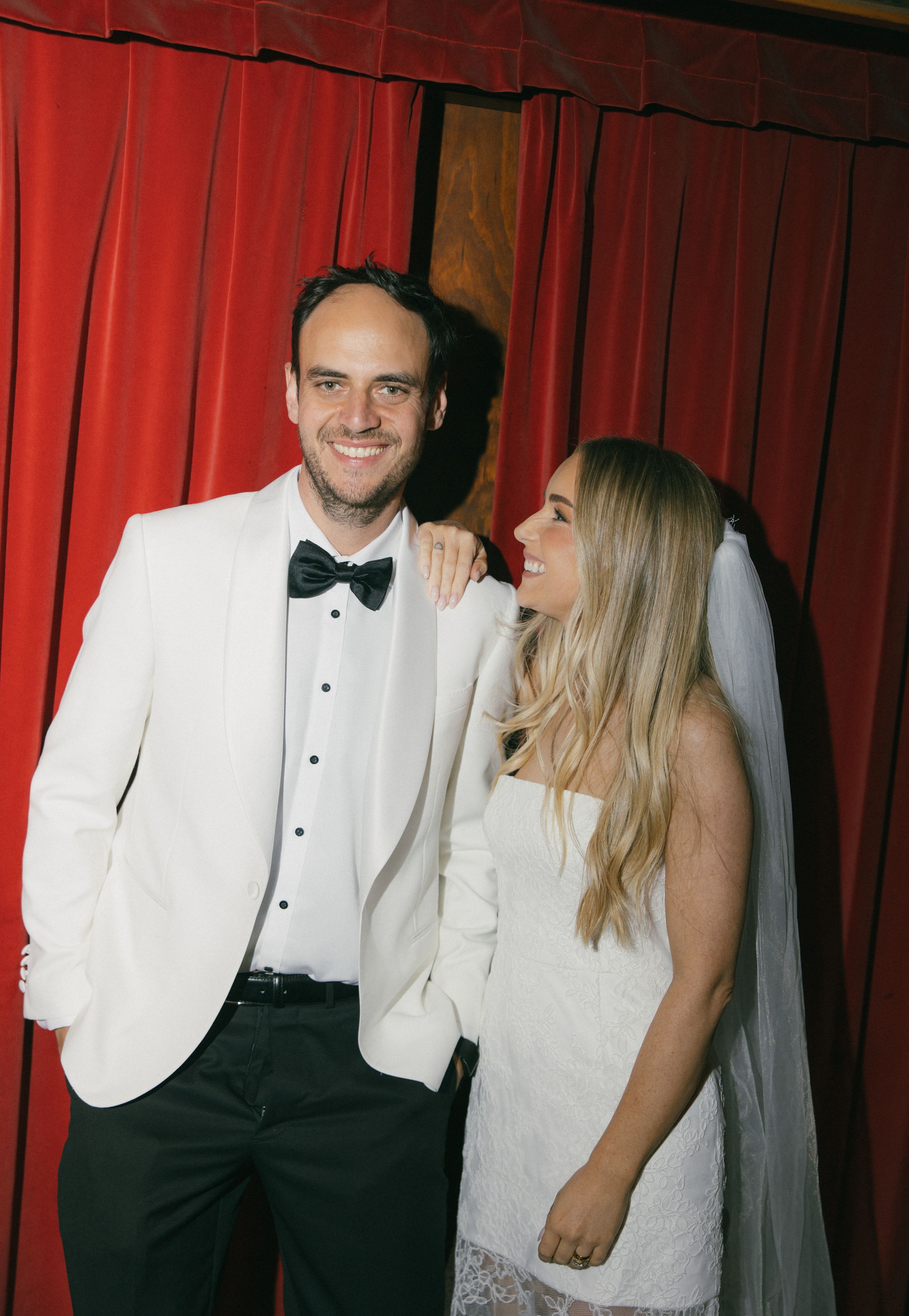 A bride and groom smile together in front of red curtains, he wears a white tuxedo with bow tie, she wears a veil.