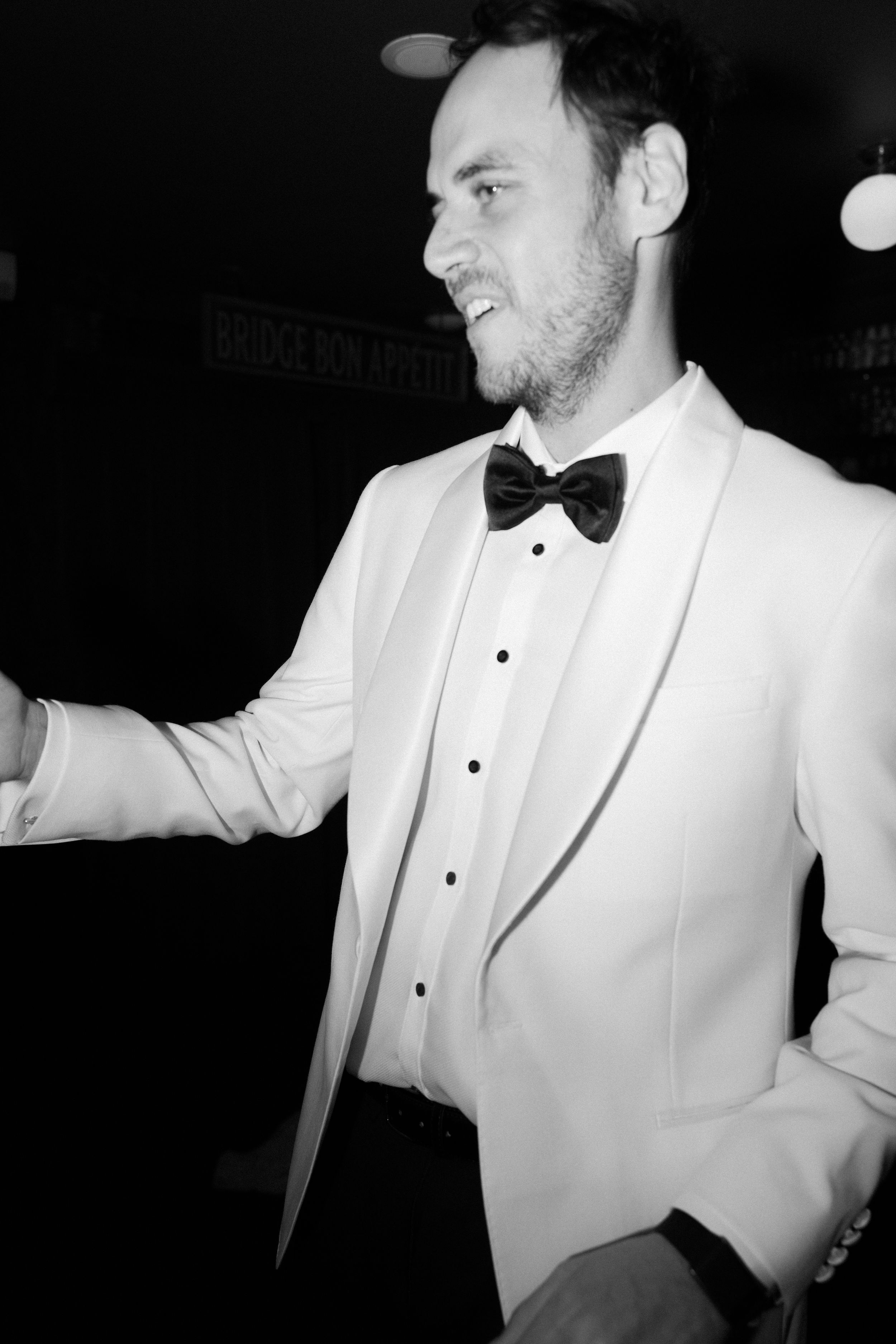 Black and white photo of a smiling man in a white tuxedo jacket and black bow tie at a formal event.
