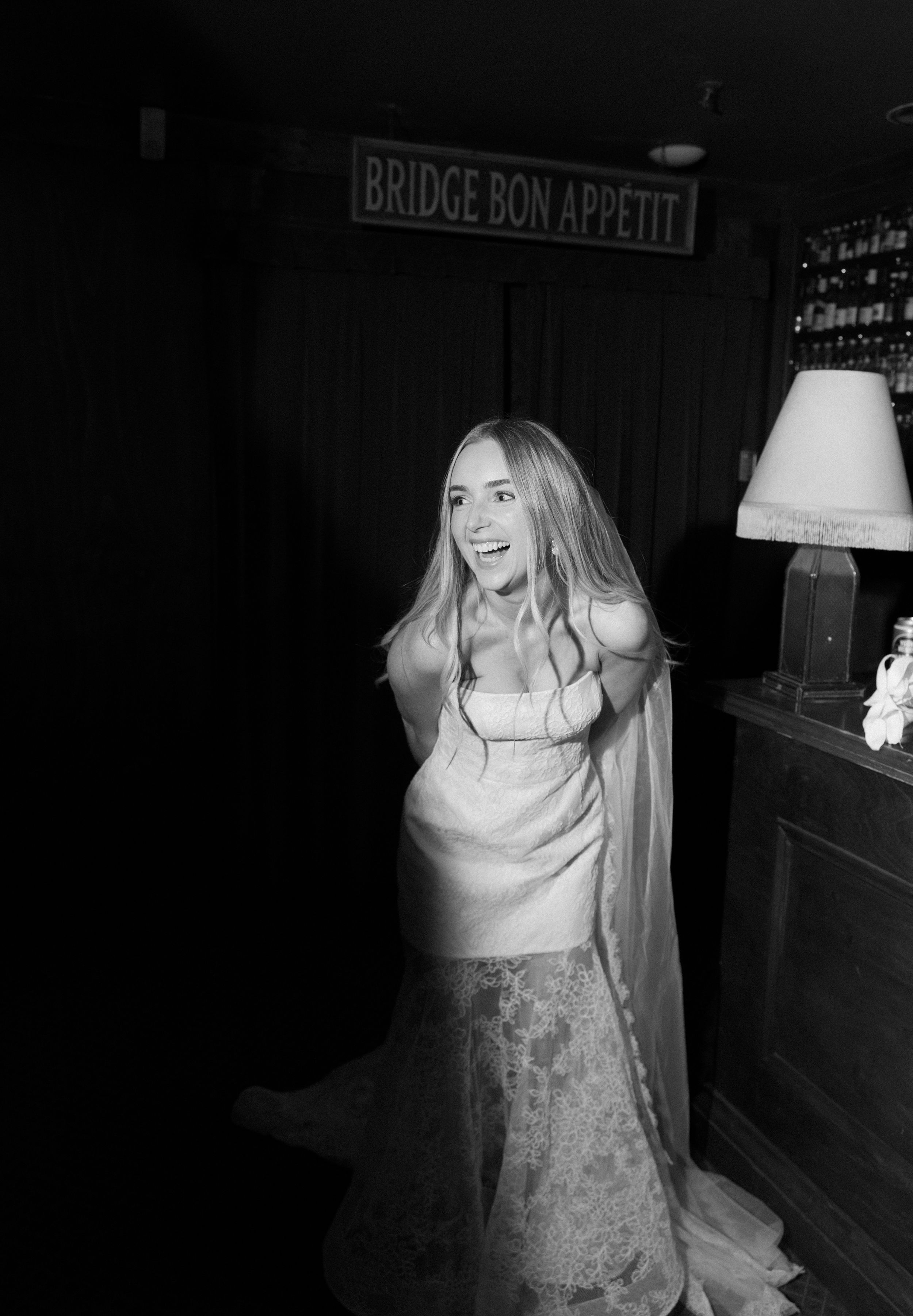 Black and white photo of a smiling blonde woman in an elegant dress near a lamp and Bridge Bon Appetit sign.