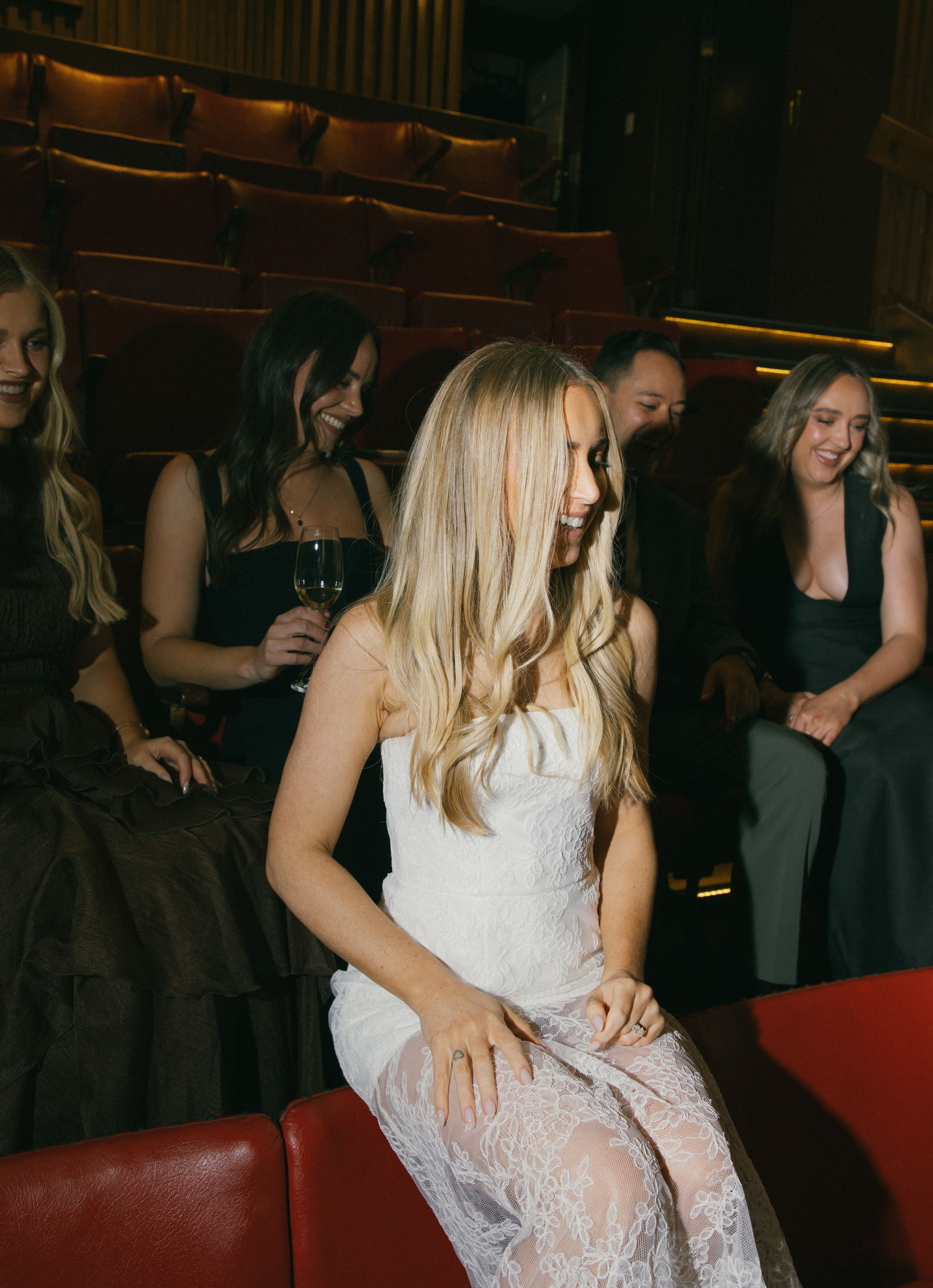 Blonde woman in white lace dress smiling at formal event, surrounded by guests in black attire in theater seating.