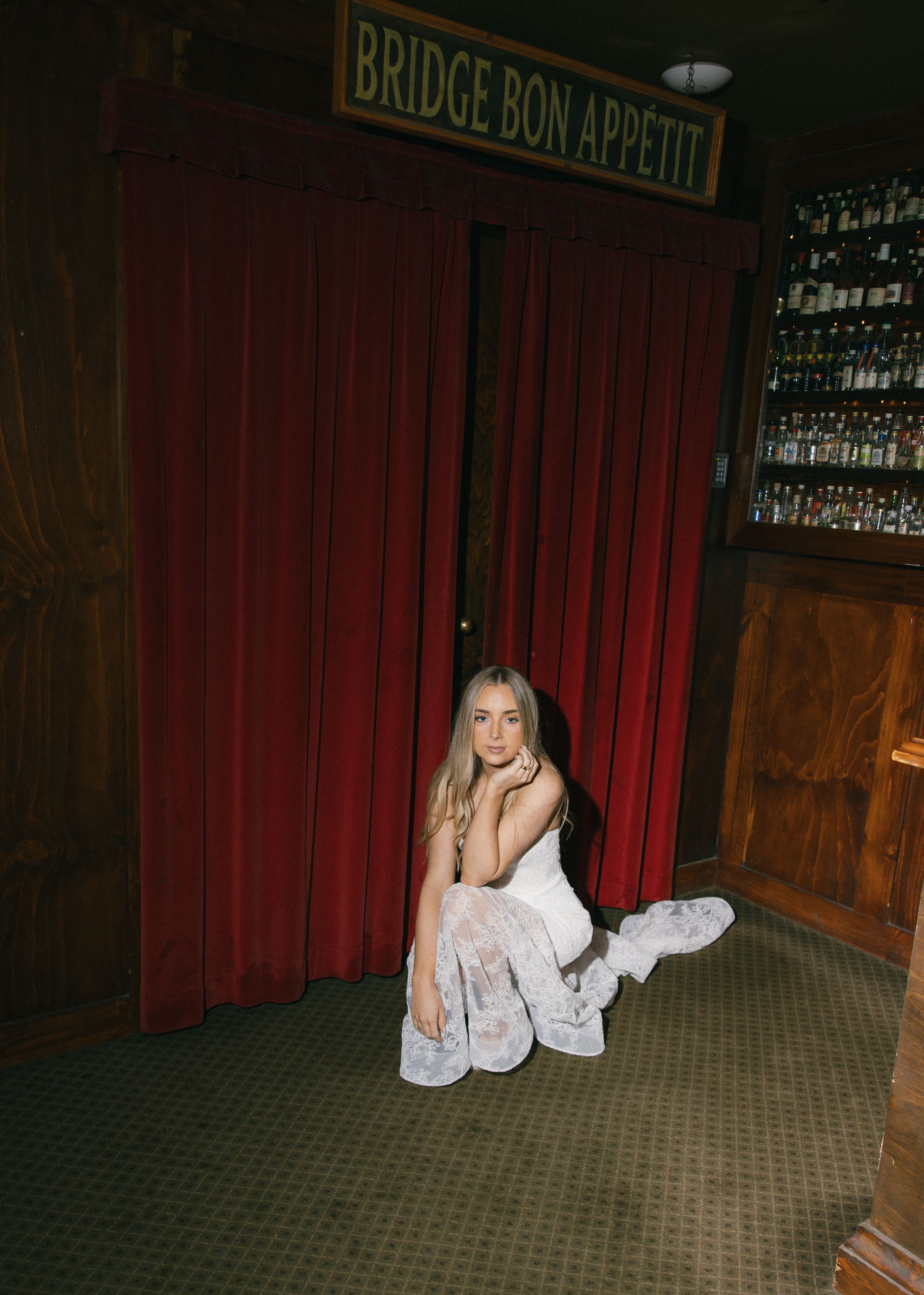 Young woman in white dress sitting on floor by red velvet curtains near a bar with a 'Bridge Bon Appetit' sign.