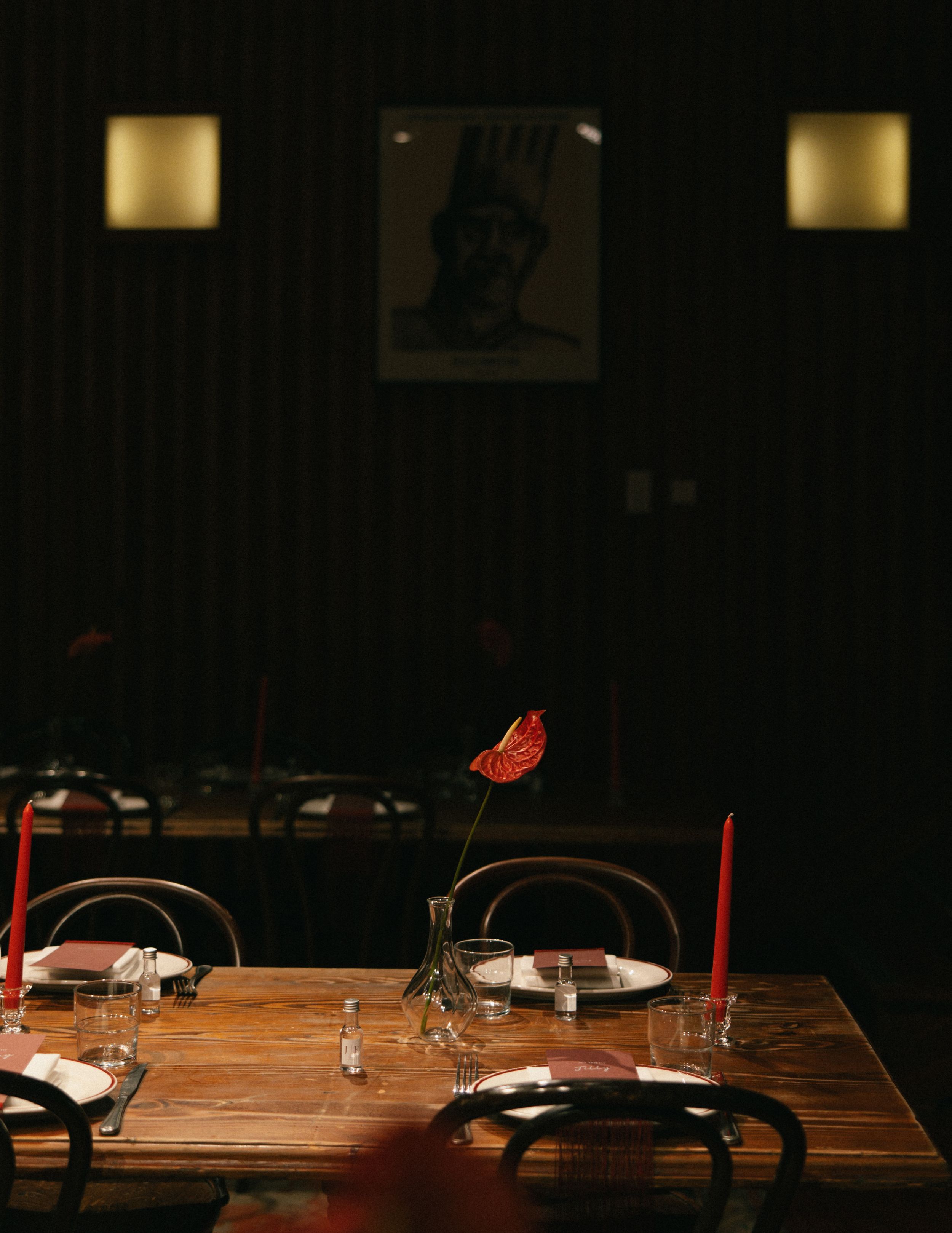 Dimly lit restaurant table set with red candles, wine glasses, and a red anthurium flower centerpiece on rustic wood.