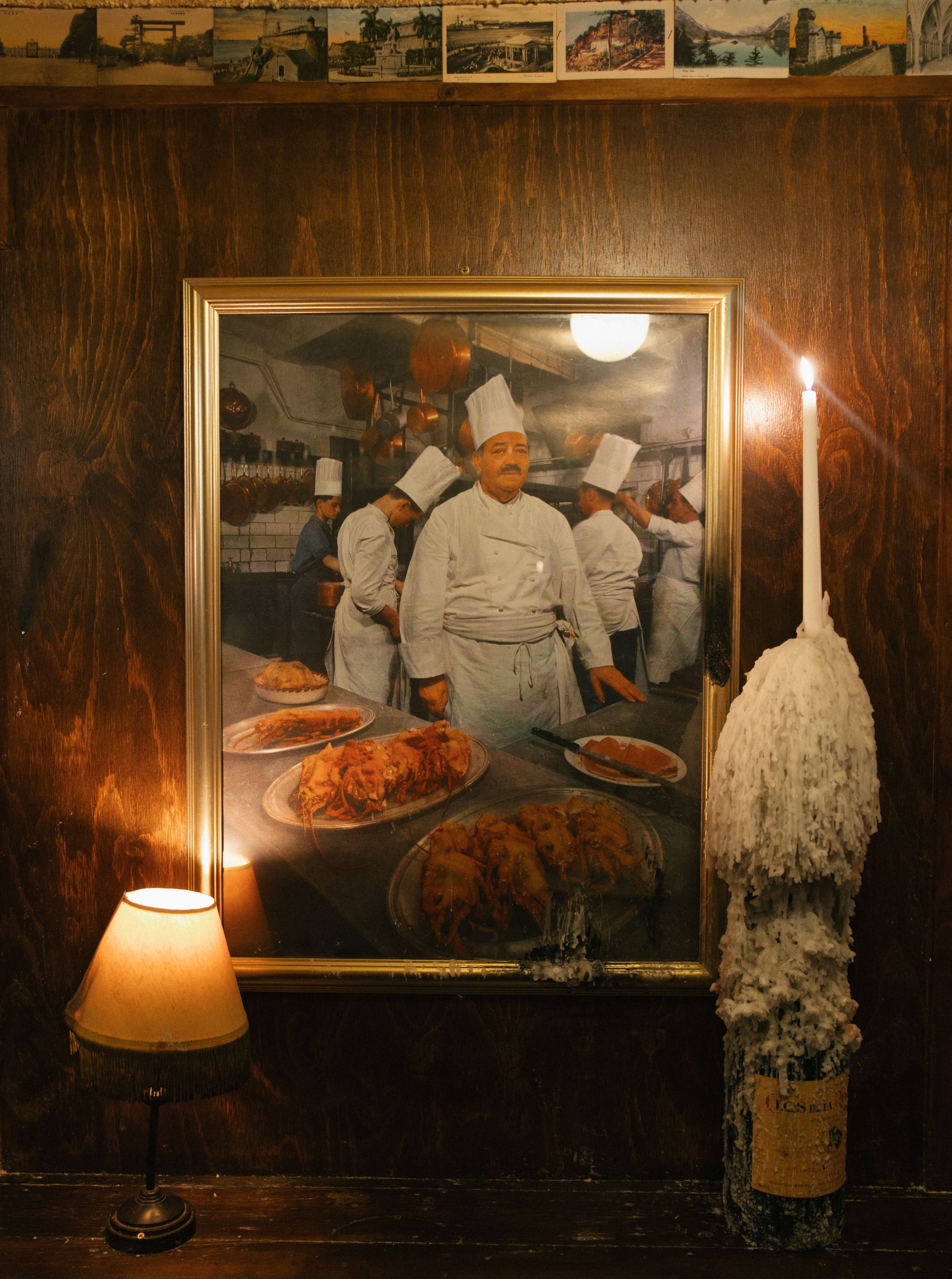 Framed photo of chefs with roasted dishes on wood-paneled wall, lit by candle and small lamp.