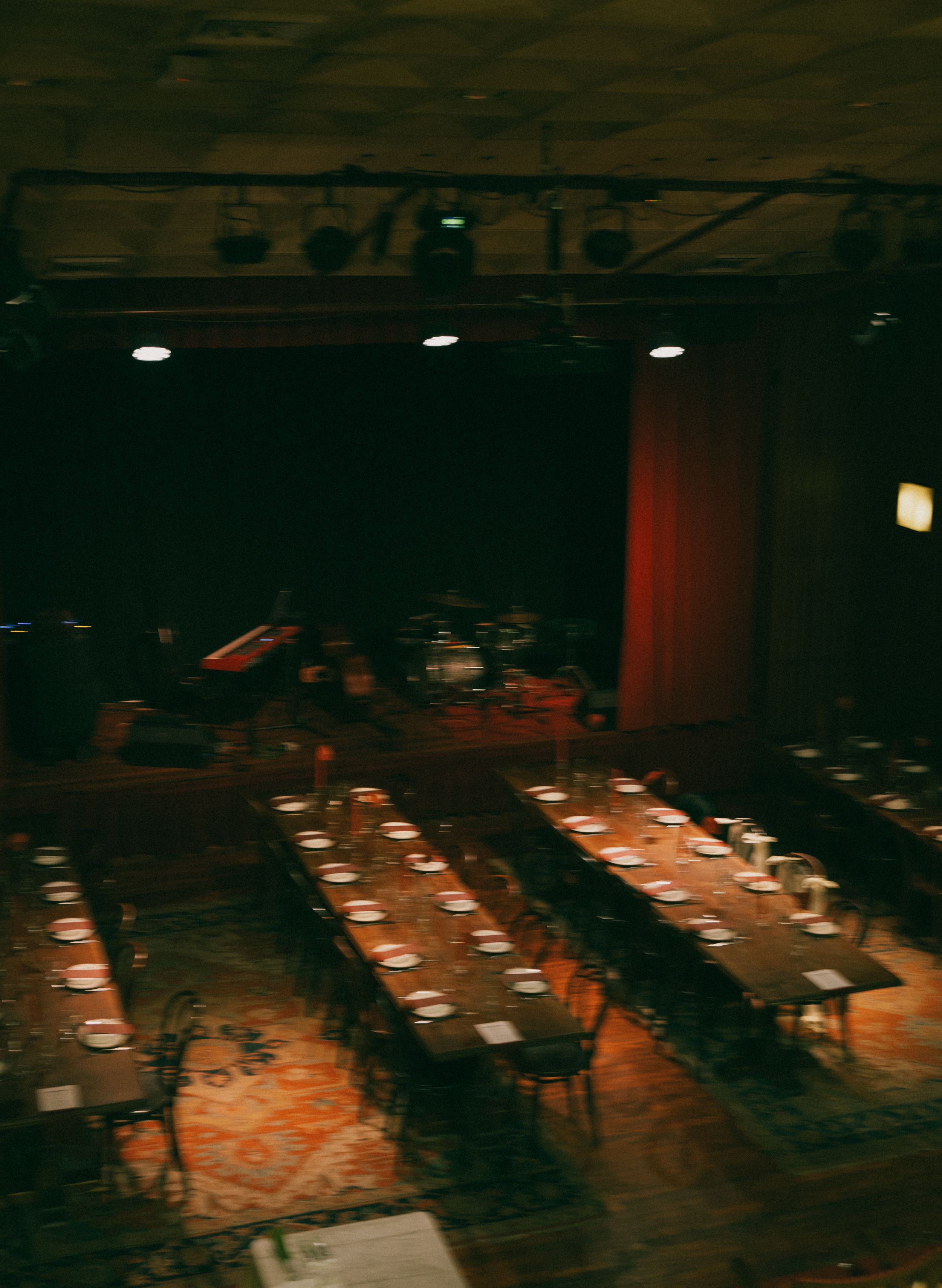 Dimly lit event venue with long banquet tables set for dining, stage with red curtain, and warm overhead lighting.