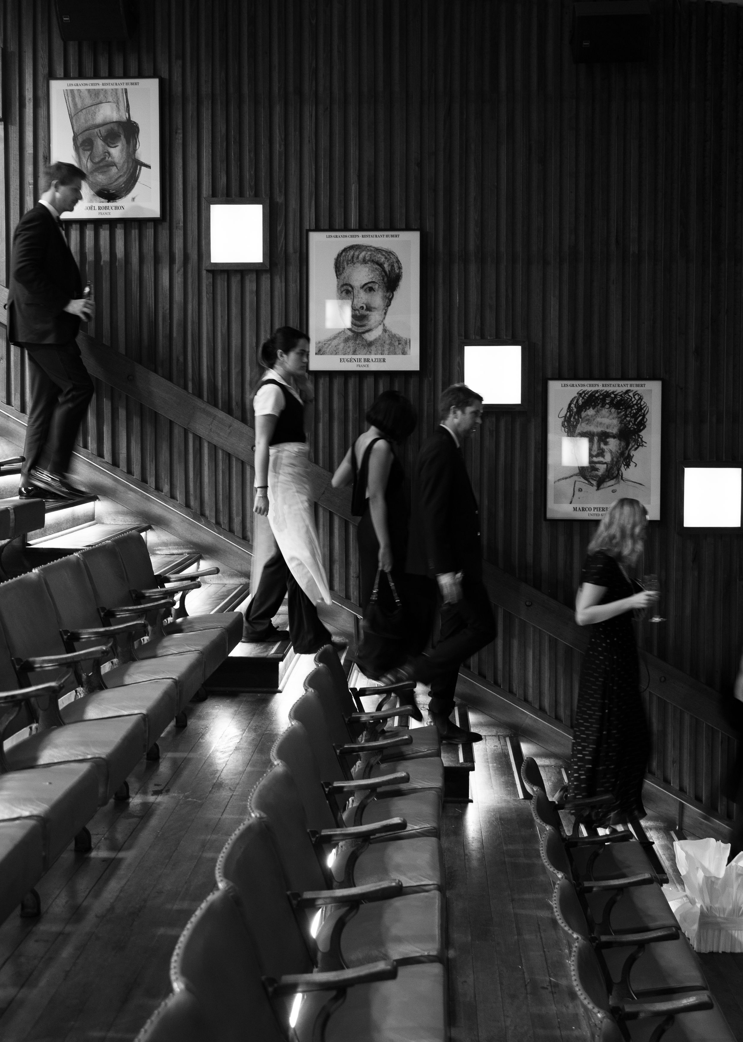 Black and white photo of people walking through a theater with portrait drawings displayed on a wood-paneled wall.