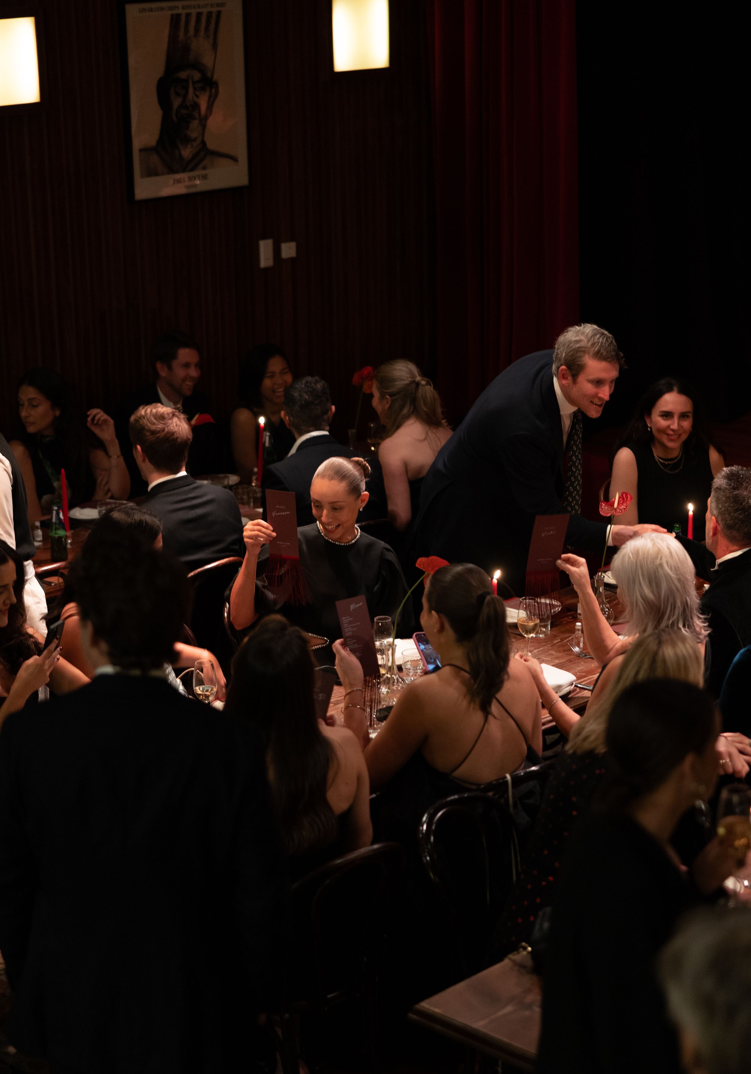 Elegant candlelit dinner party with guests in formal attire gathered around tables in a dimly lit venue.