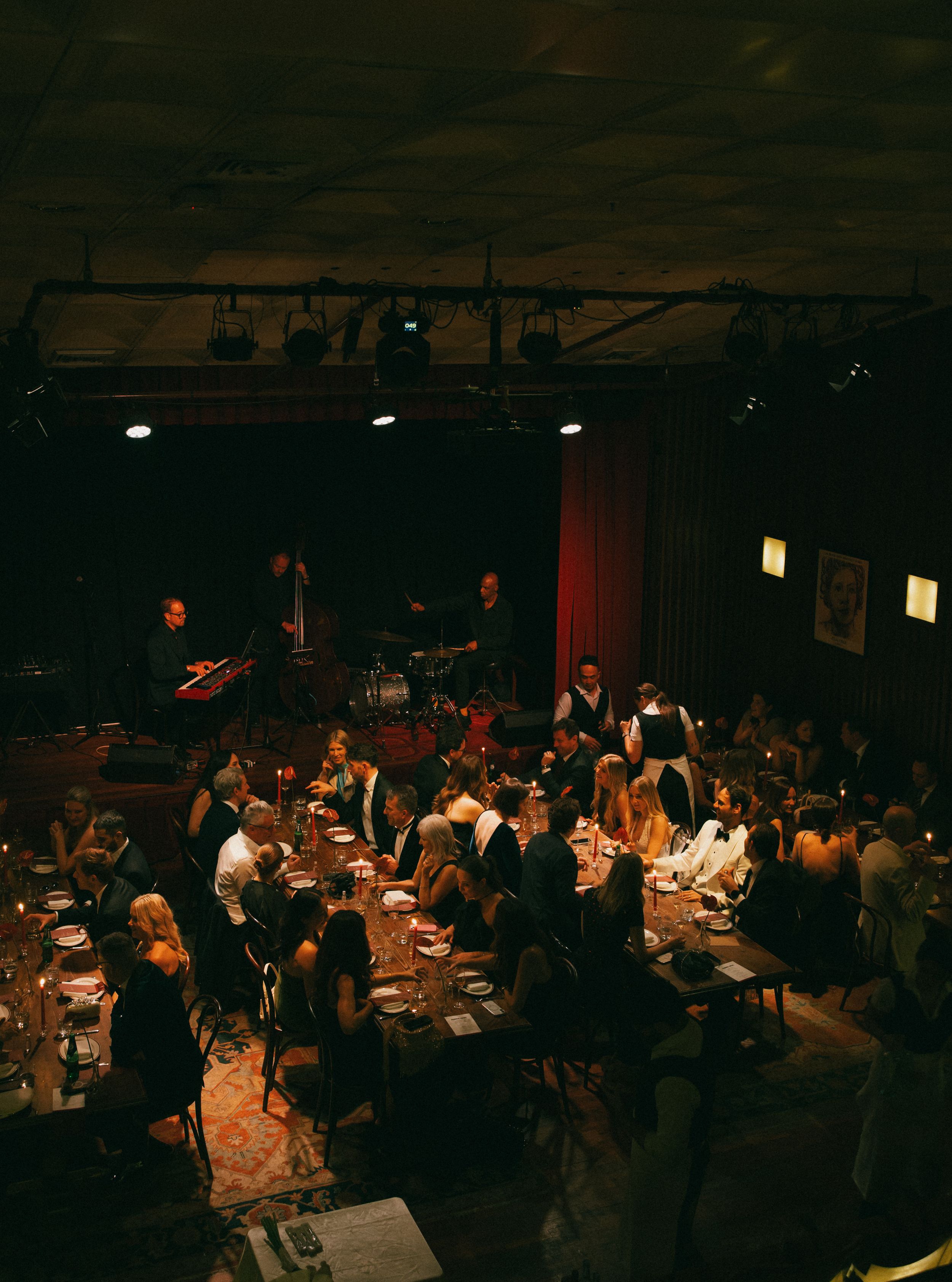 Elegant dinner event in a dimly lit venue with guests at candlelit tables and a live band performing on stage.