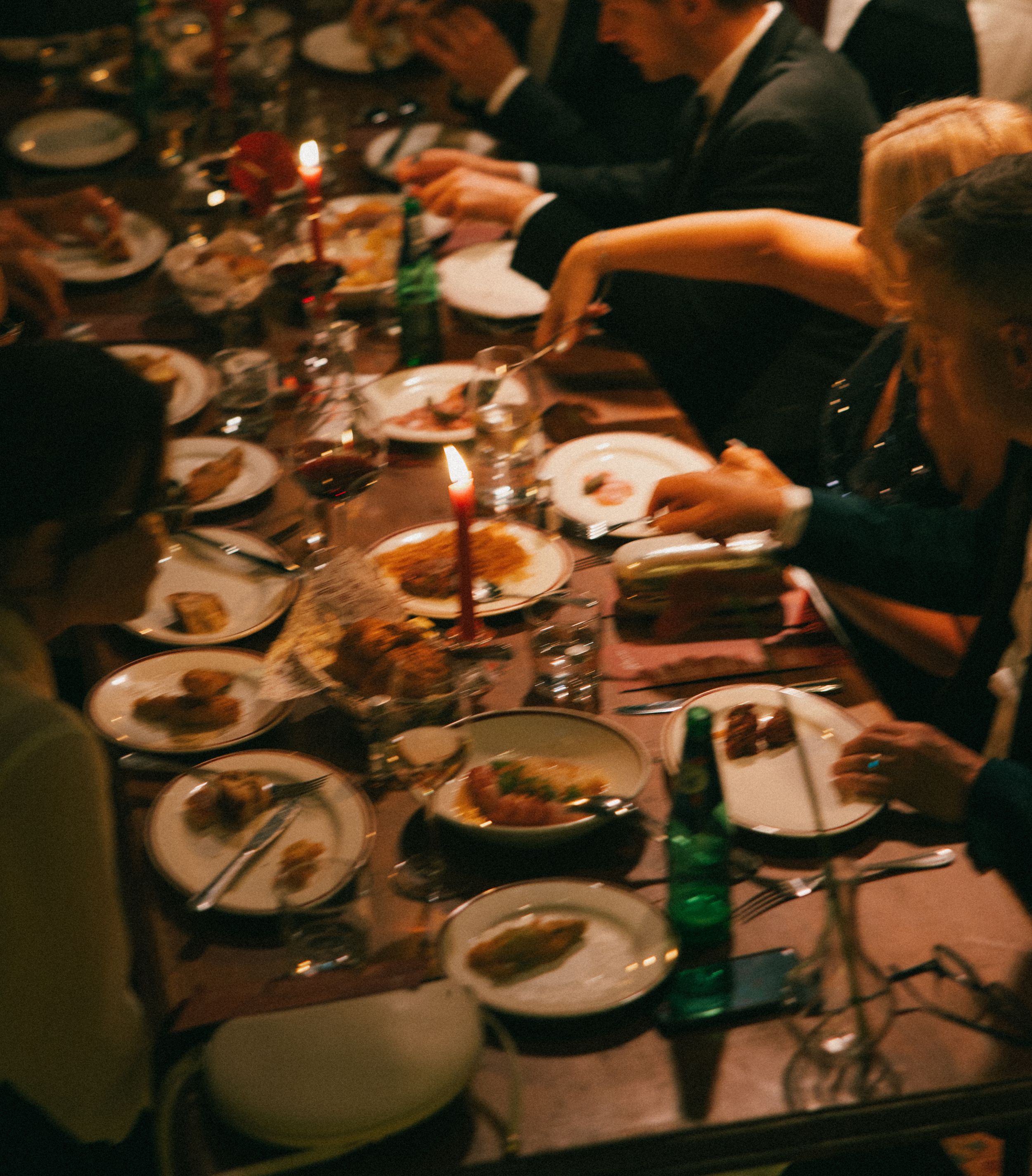 Elegant candlelit dinner party with guests dining at a long table filled with plates, glassware, and green bottles.