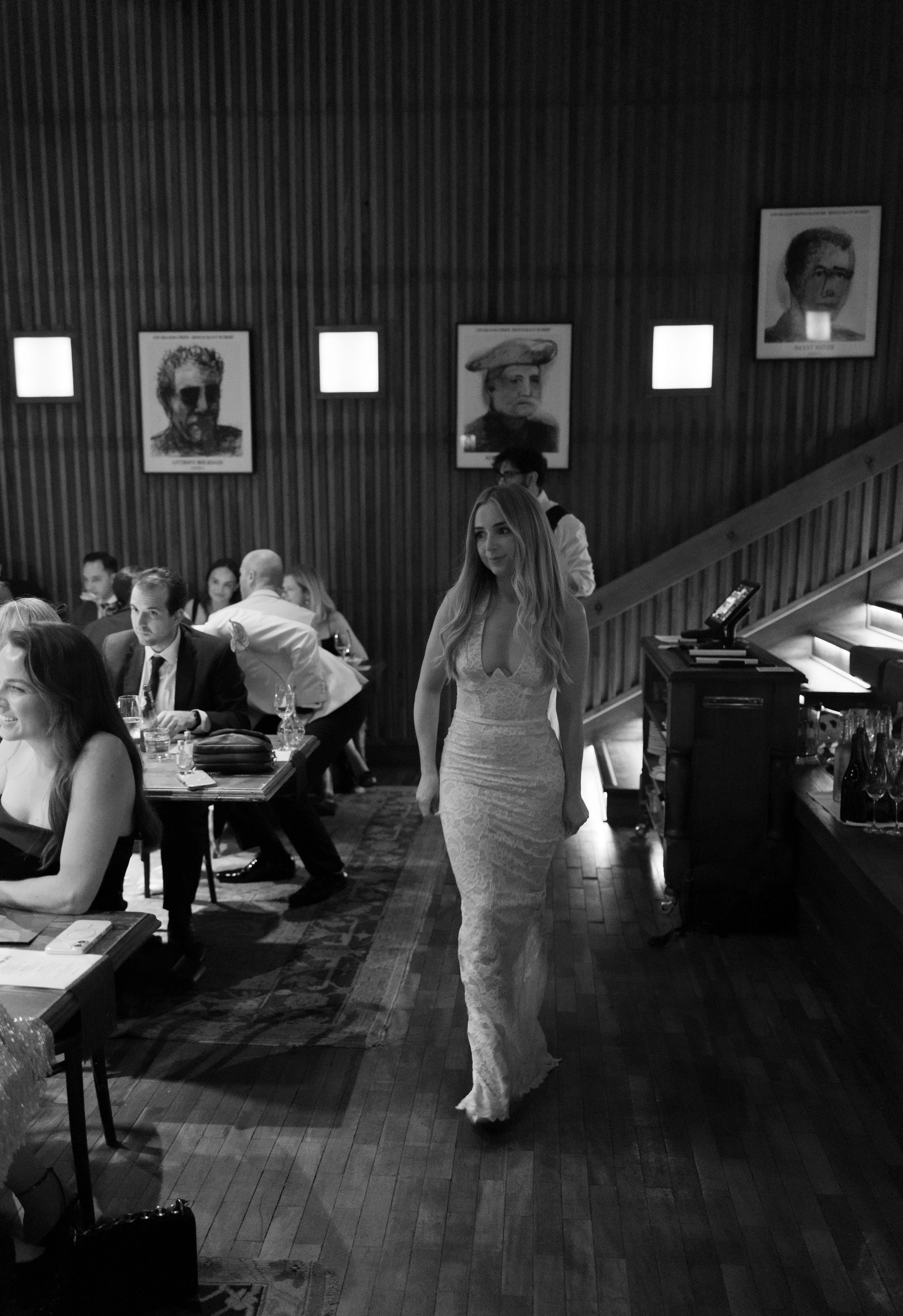 Black and white photo of an elegant woman in a lace gown walking through a upscale restaurant with portraits on walls.