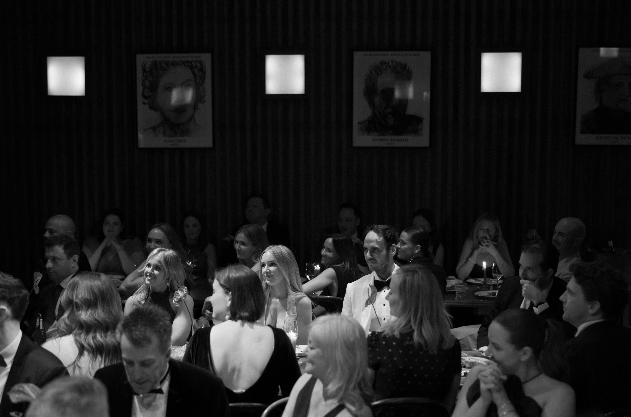 Black and white photo of elegantly dressed guests mingling at a formal gala dinner event in a dimly lit venue.