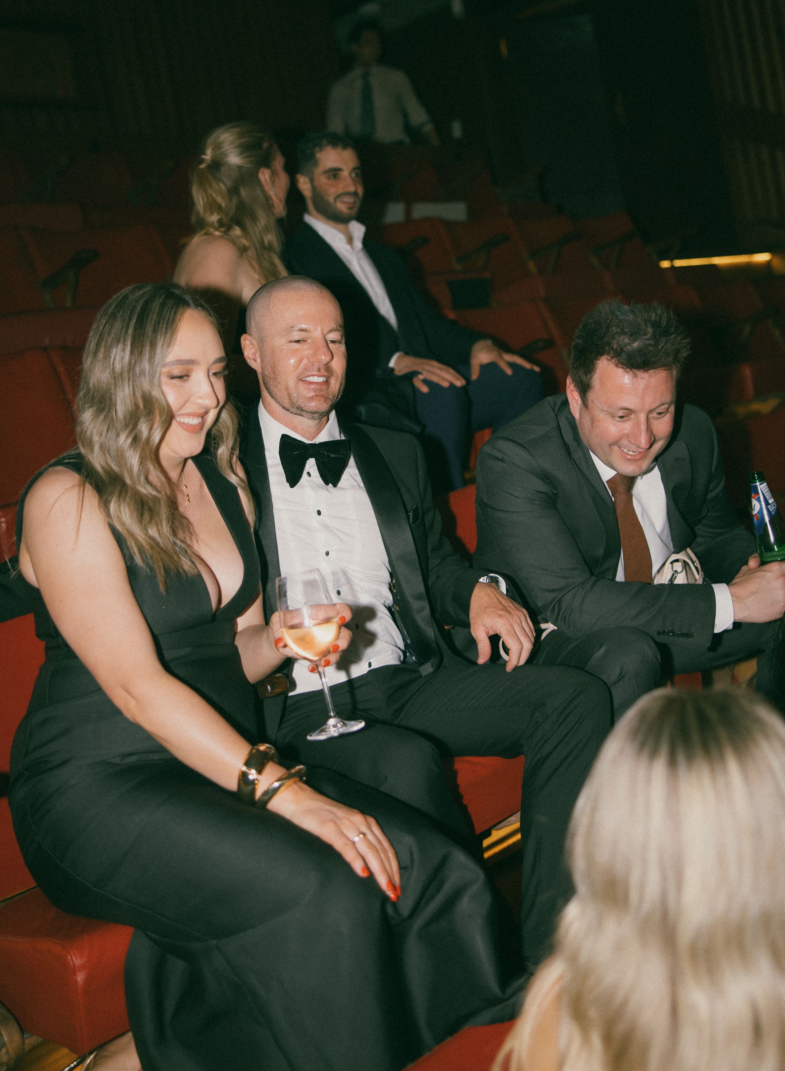 Elegantly dressed guests in black tie attire laughing and socializing in red theater seats at a formal event.